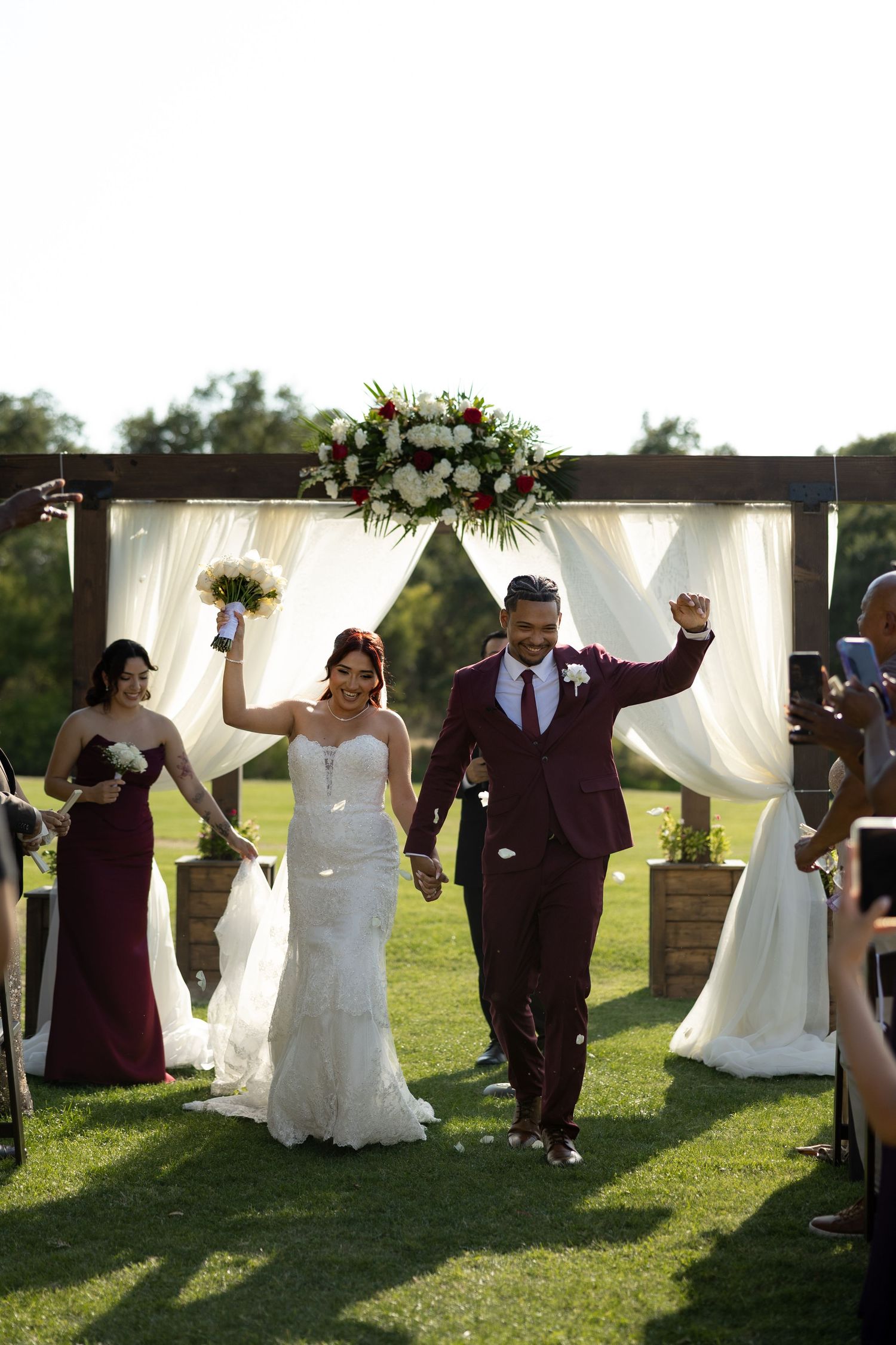 Joyful newlyweds celebrate recessional at outdoor wedding ceremony with floral arch, bride in white gown, groom in burgundy suit.