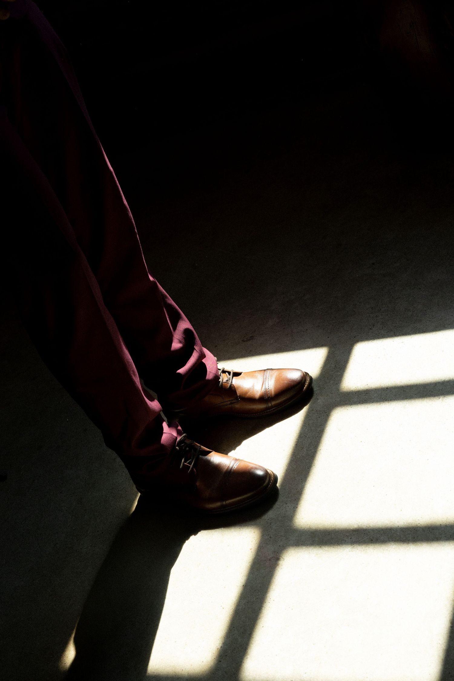 Close-up of men's brown leather dress shoes with burgundy trousers standing in dramatic window light on dark floor.