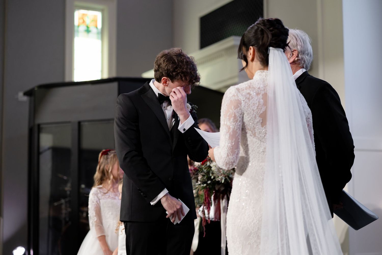 Emotional groom wiping tears as bride approaches at elegant church wedding ceremony with flower girl.