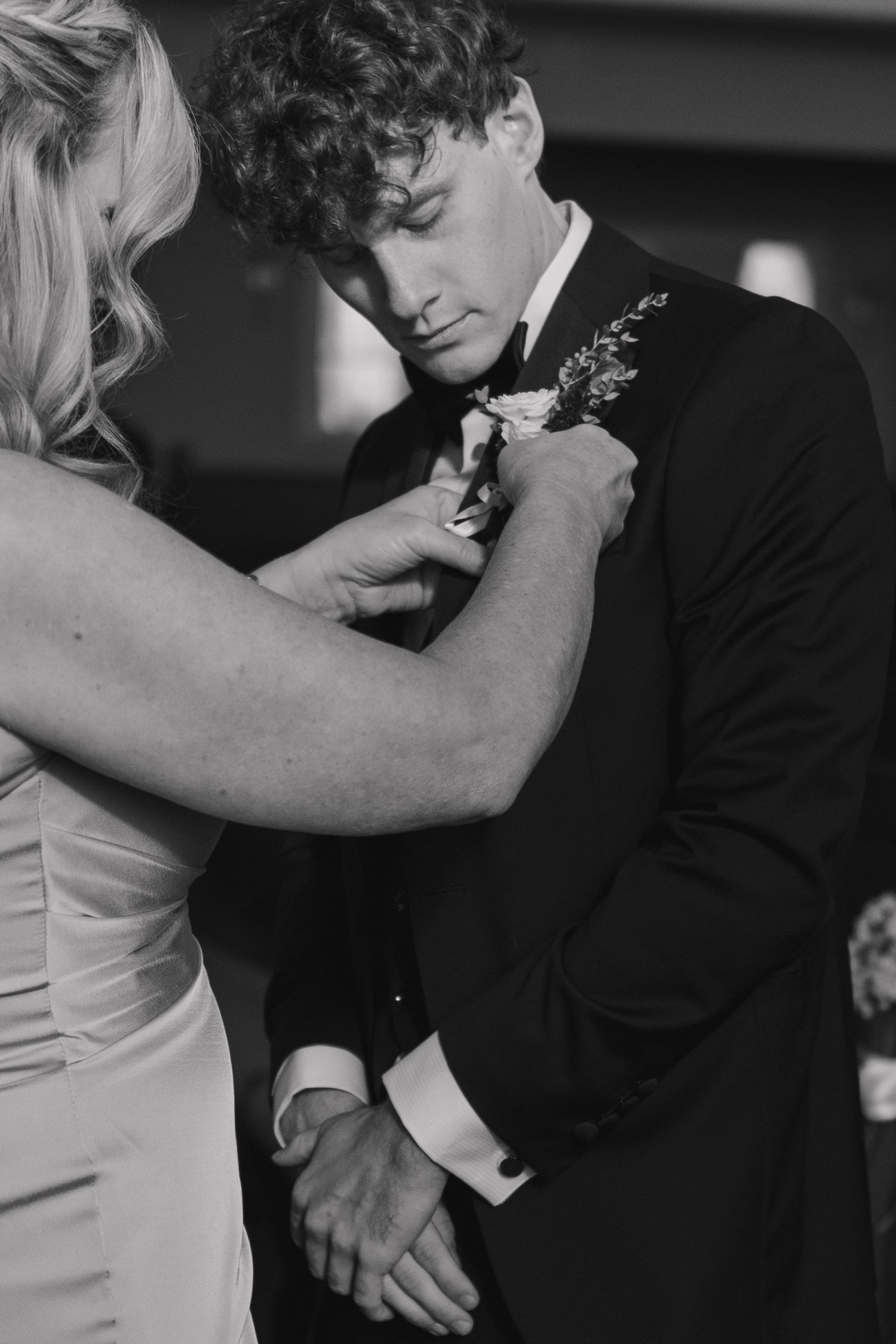 Black and white photo of a woman pinning a boutonniere on a groom in a black tuxedo.