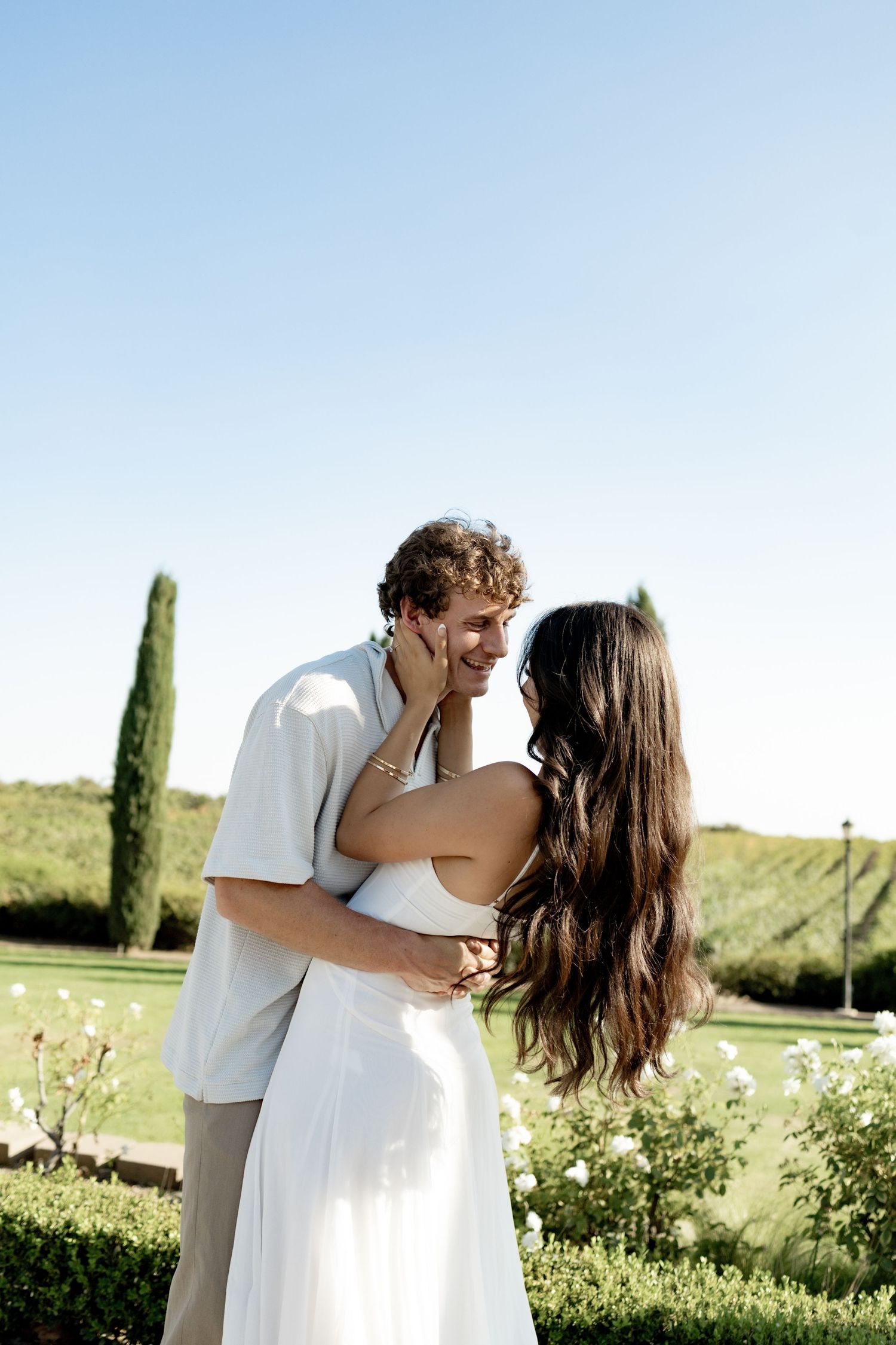 A romantic outdoor photo sequence showing a couple embracing in a sunny garden setting with cypress trees.
