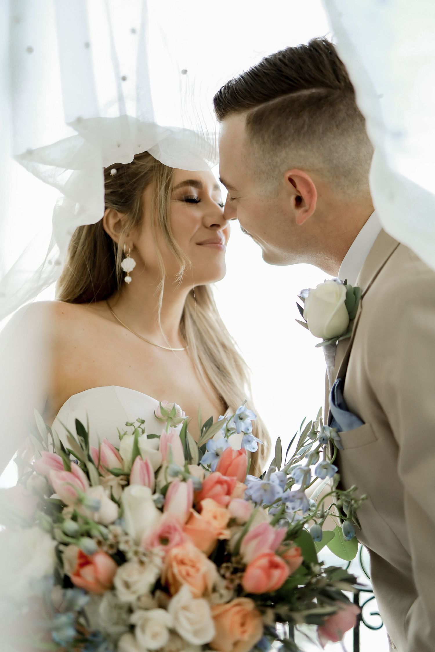 Wedding couple posing intimately with colorful floral bouquet against white curtain backdrop.