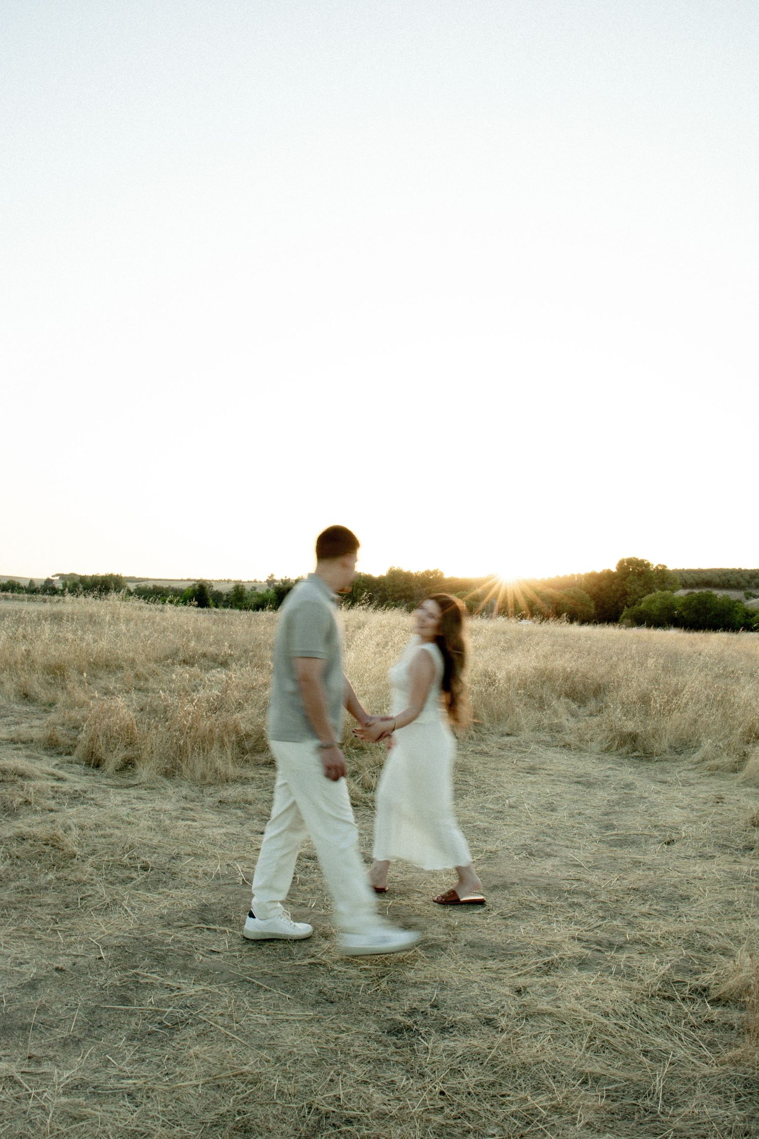A couple dances together in a golden field at sunset while wearing white clothing.