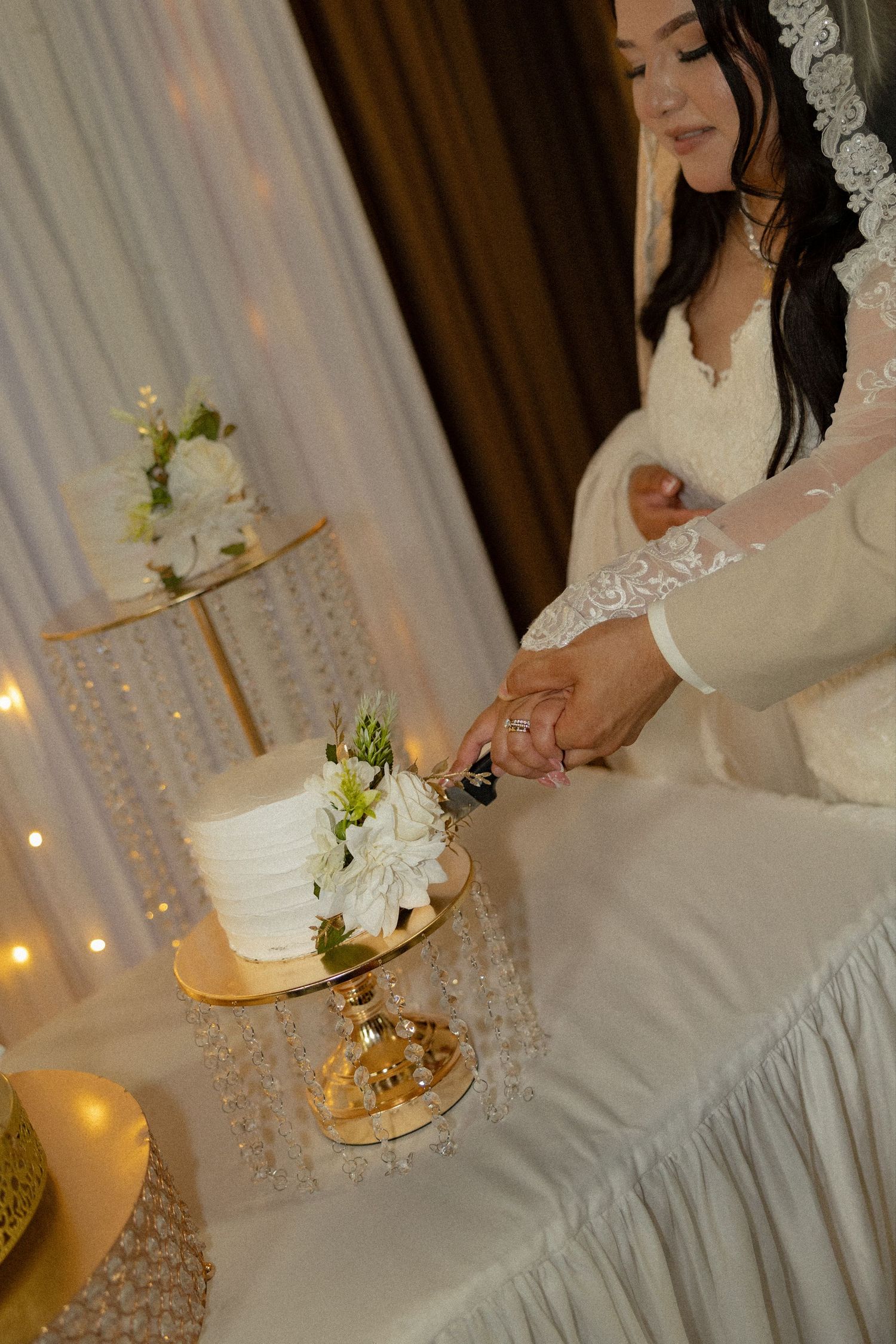 Bride in lace wedding dress cutting a white floral cake on a gold crystal stand at a wedding reception.