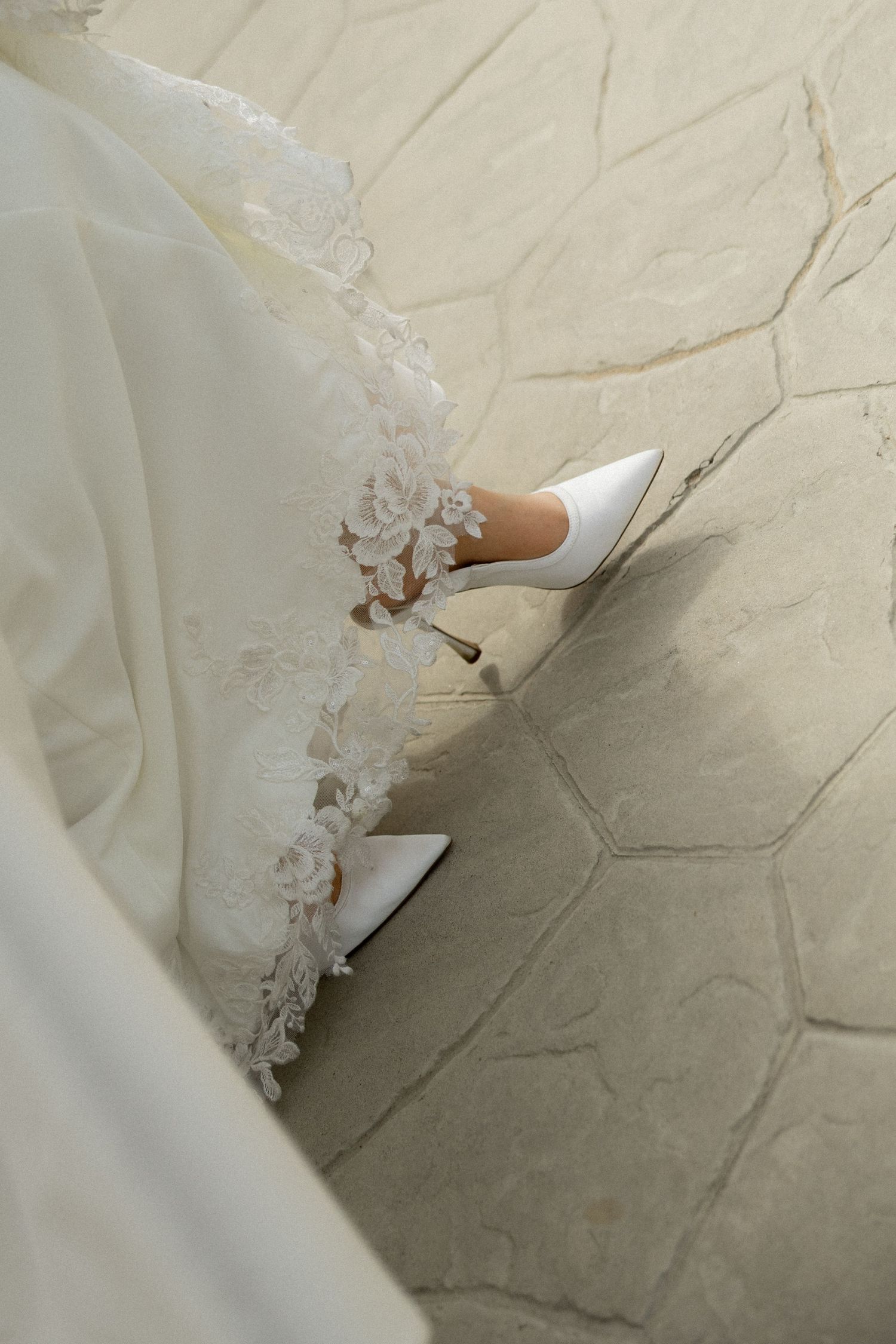 Bride wearing white stiletto heels with floral ankle details and lace-trimmed wedding gown on hexagonal stone floor.