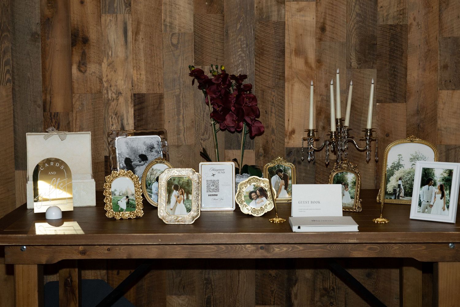 Elegant wedding memory table with gold frames, family photos, candelabra, and dark red florals on a rustic wood backdrop.