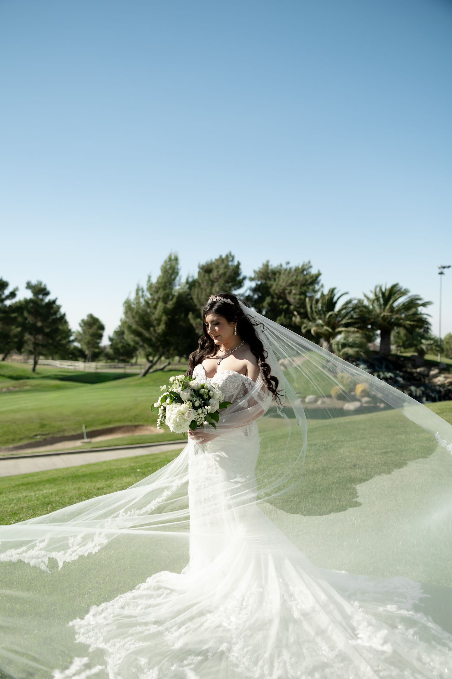 Bride in white lace mermaid gown with flowing veil holding bouquet on green golf course under clear blue sky.
