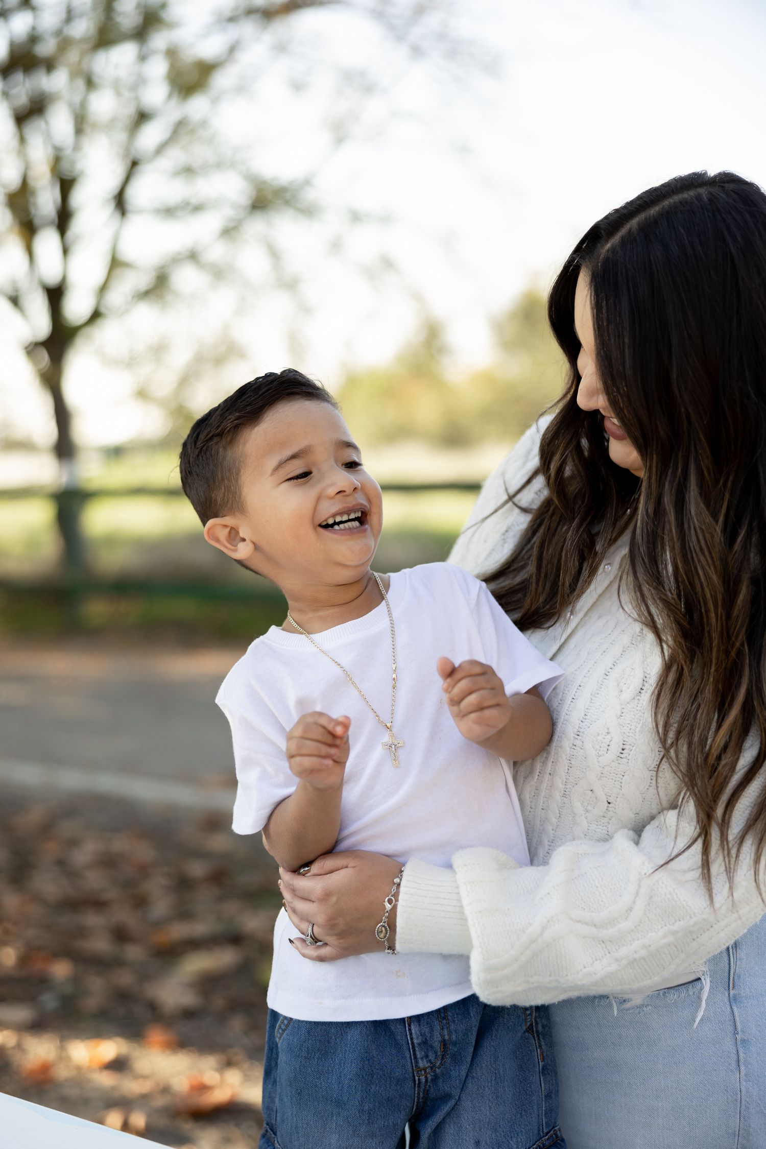 A joyful child in a white t-shirt and jeans laughs while being embraced by someone in a white sweater outdoors.