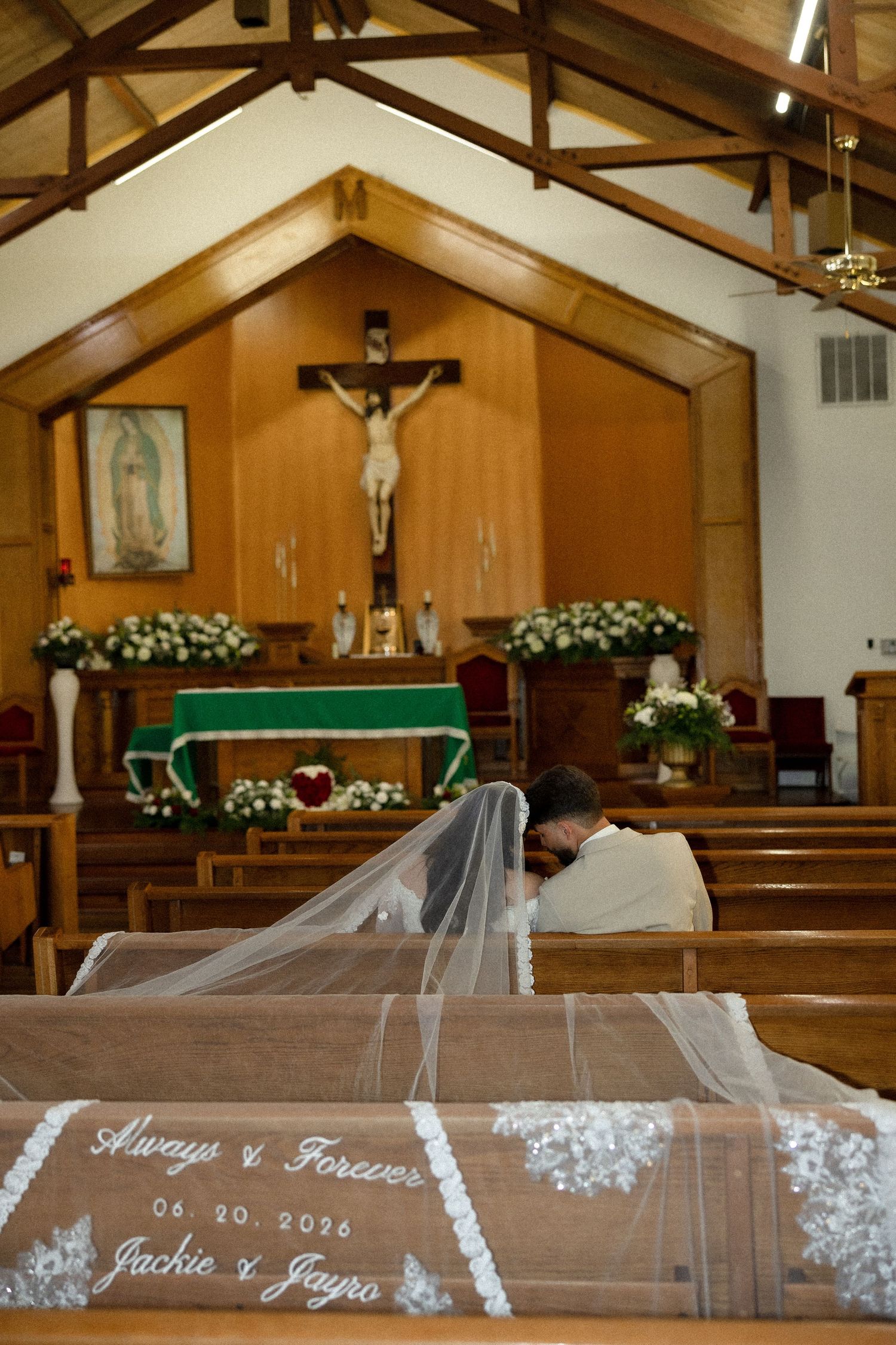 Bride and groom kneeling in prayer at a Catholic church altar, wedding veil embroidered with Always & Forever 06.20.2026 Jackie & Jayro.