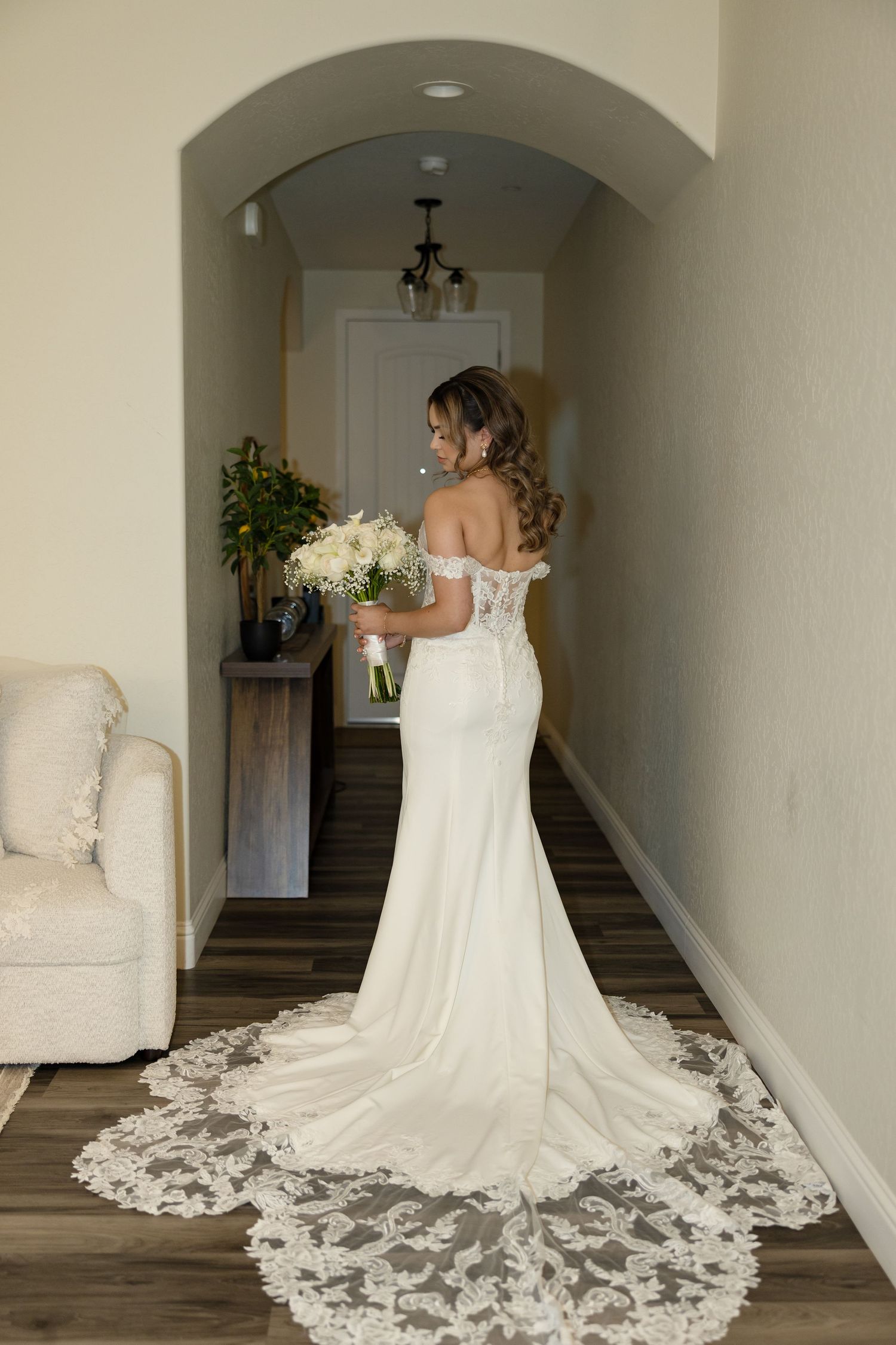 Bride in elegant off-shoulder lace wedding gown with cathedral train holds white bouquet in a hallway.