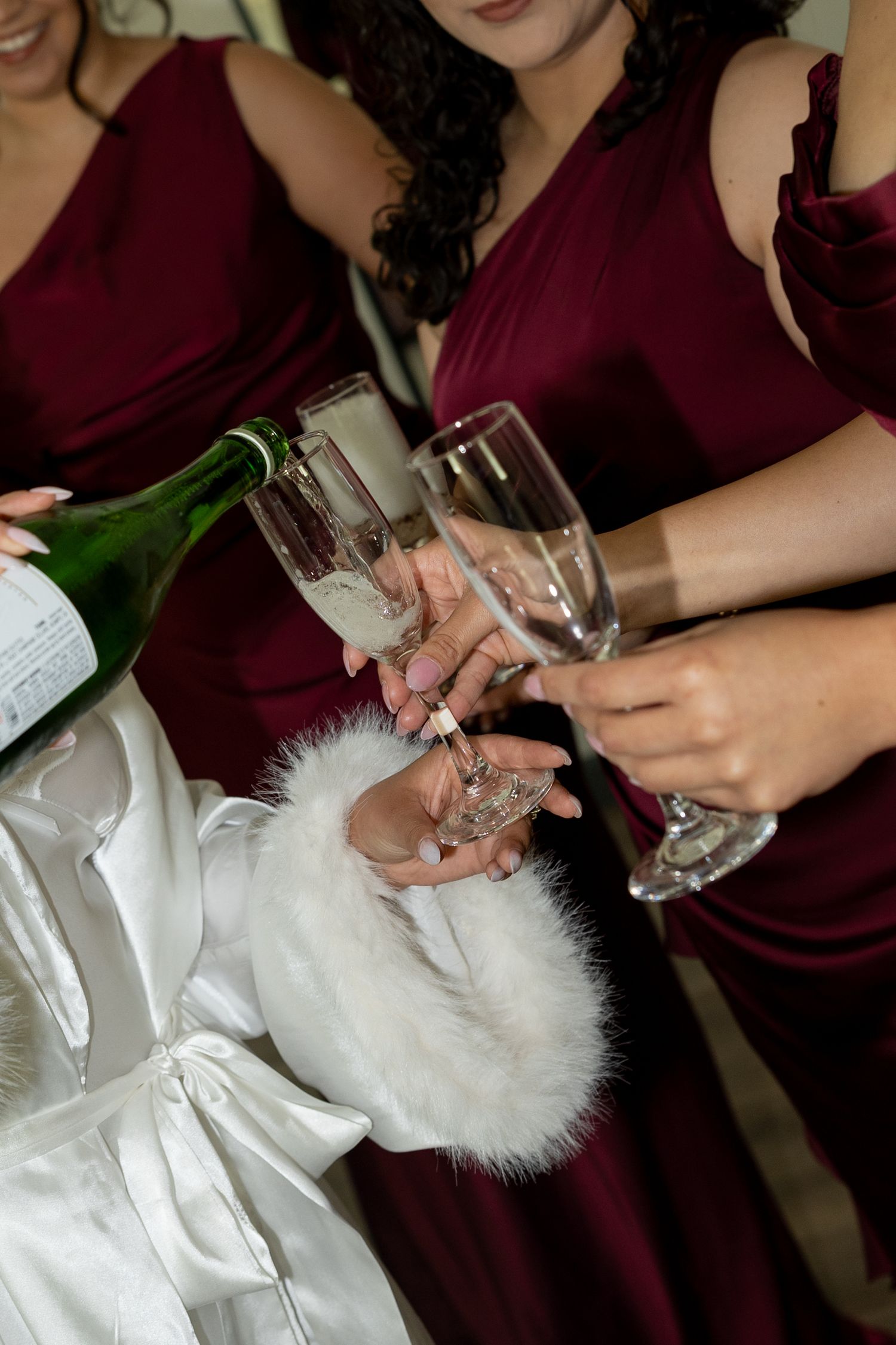 Bridesmaids in burgundy dresses toasting with champagne flutes while champagne is poured at a wedding celebration.
