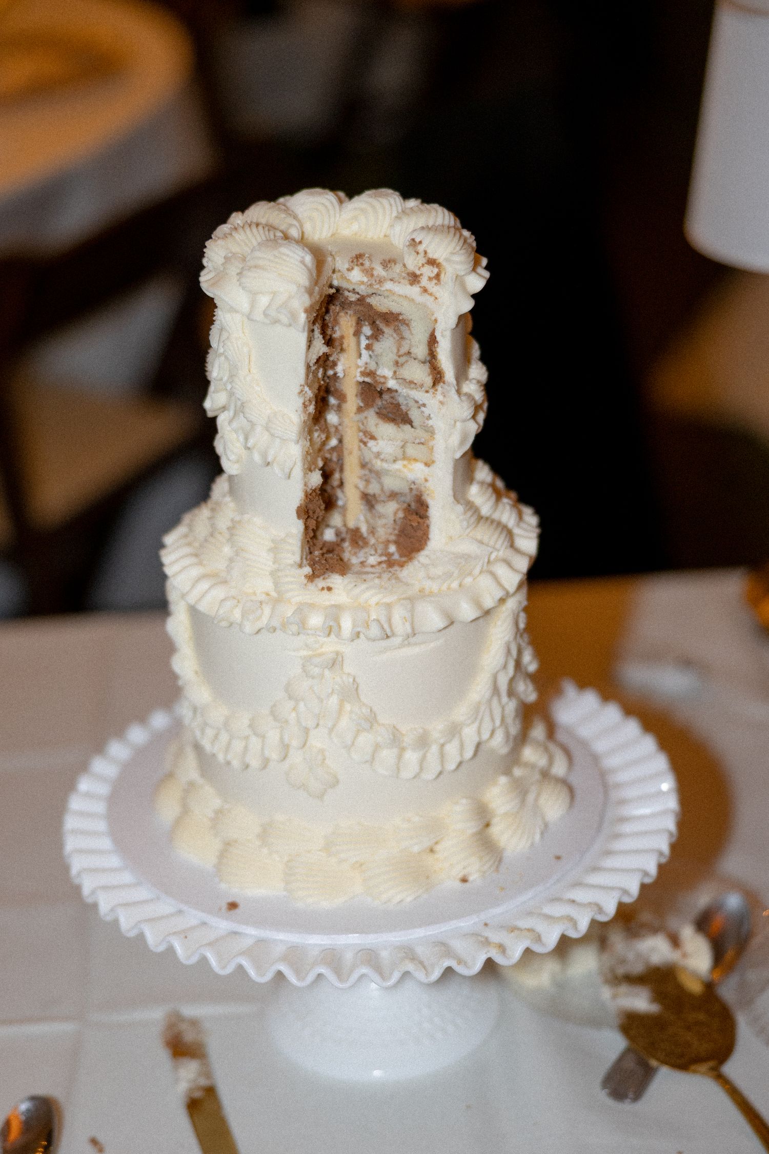 A two-tier white wedding cake with decorative frosting, partially cut to reveal chocolate interior, displayed on a white cake stand.