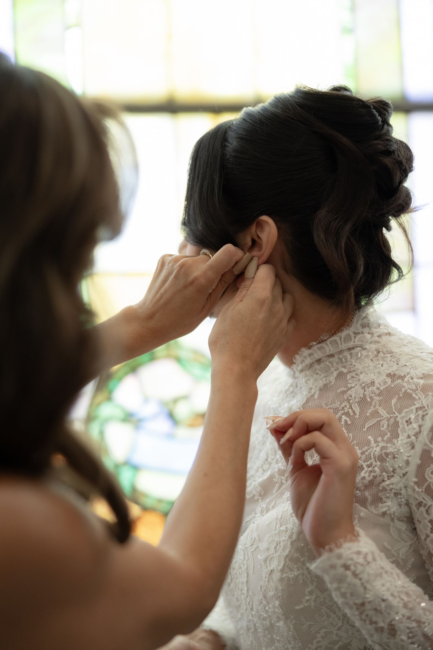 Bridesmaid helping bride put on earrings before wedding ceremony near stained glass window.