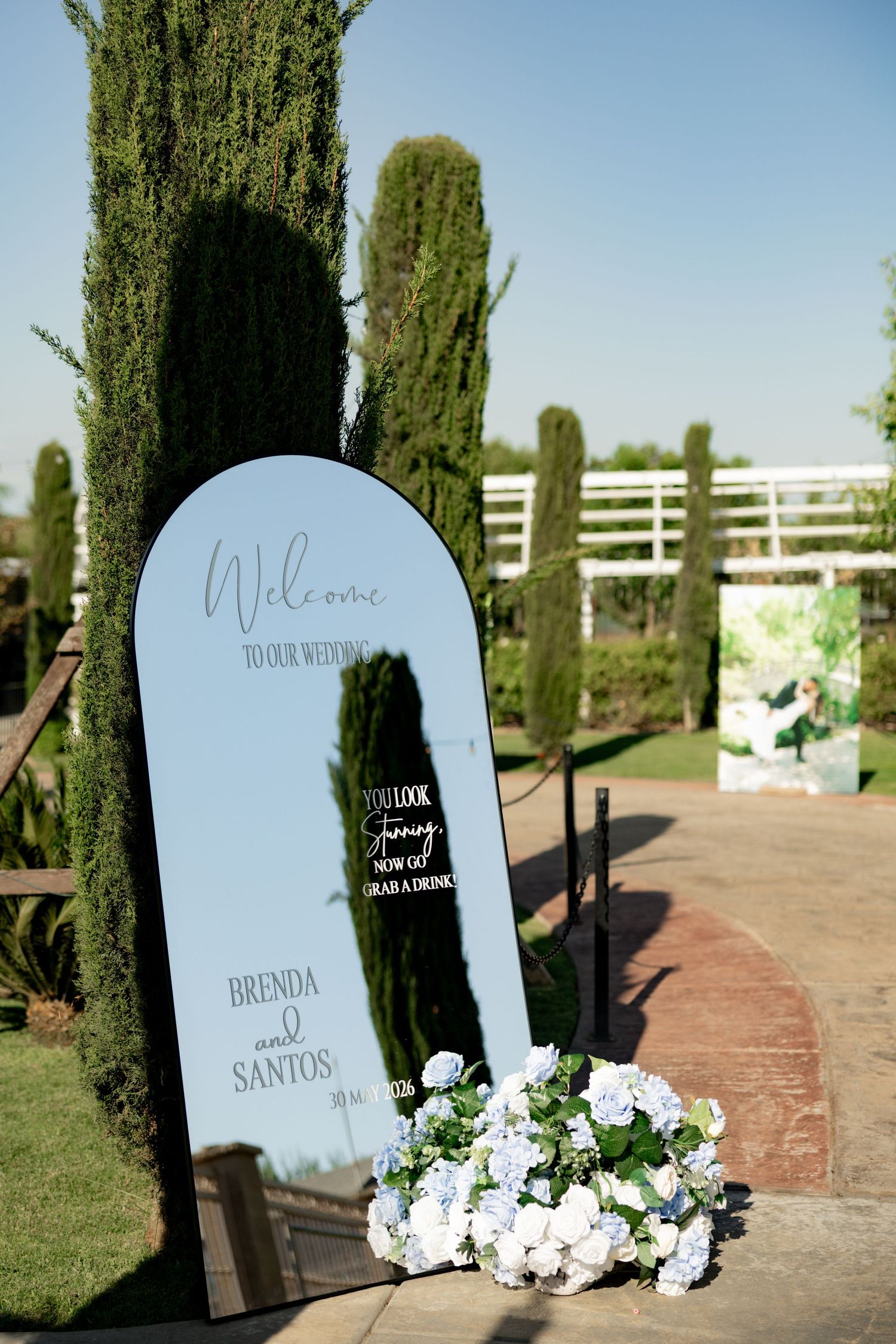 Elegant arched wedding welcome mirror sign for Brenda and Santos, adorned with white hydrangeas, set in a lush garden pathway.