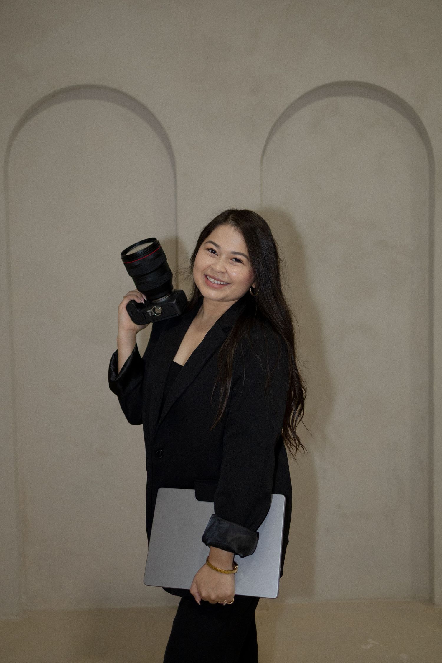 Professional photographer in black attire poses with camera equipment against neutral backdrop in studio setting.