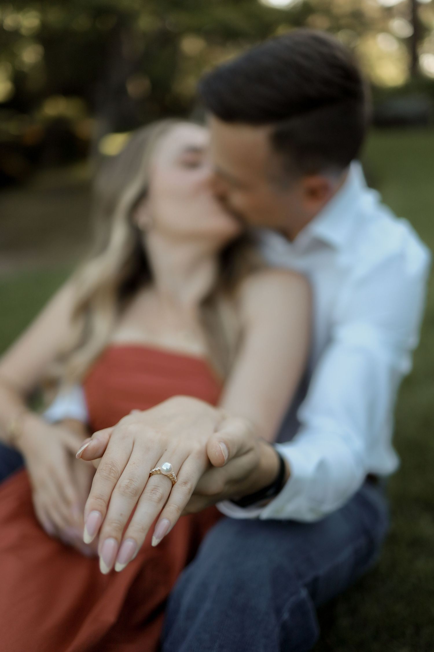 A close-up of hands showing off a sparkling engagement ring against a red dress in an outdoor setting.