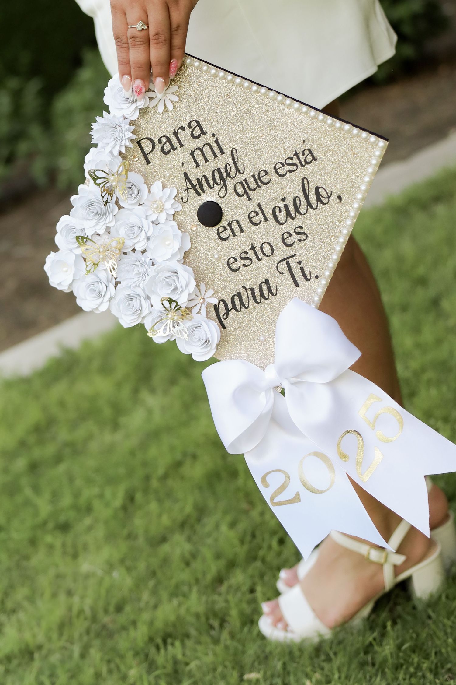 Decorated graduation cap with white flowers and Spanish text shown in detail against grass background.