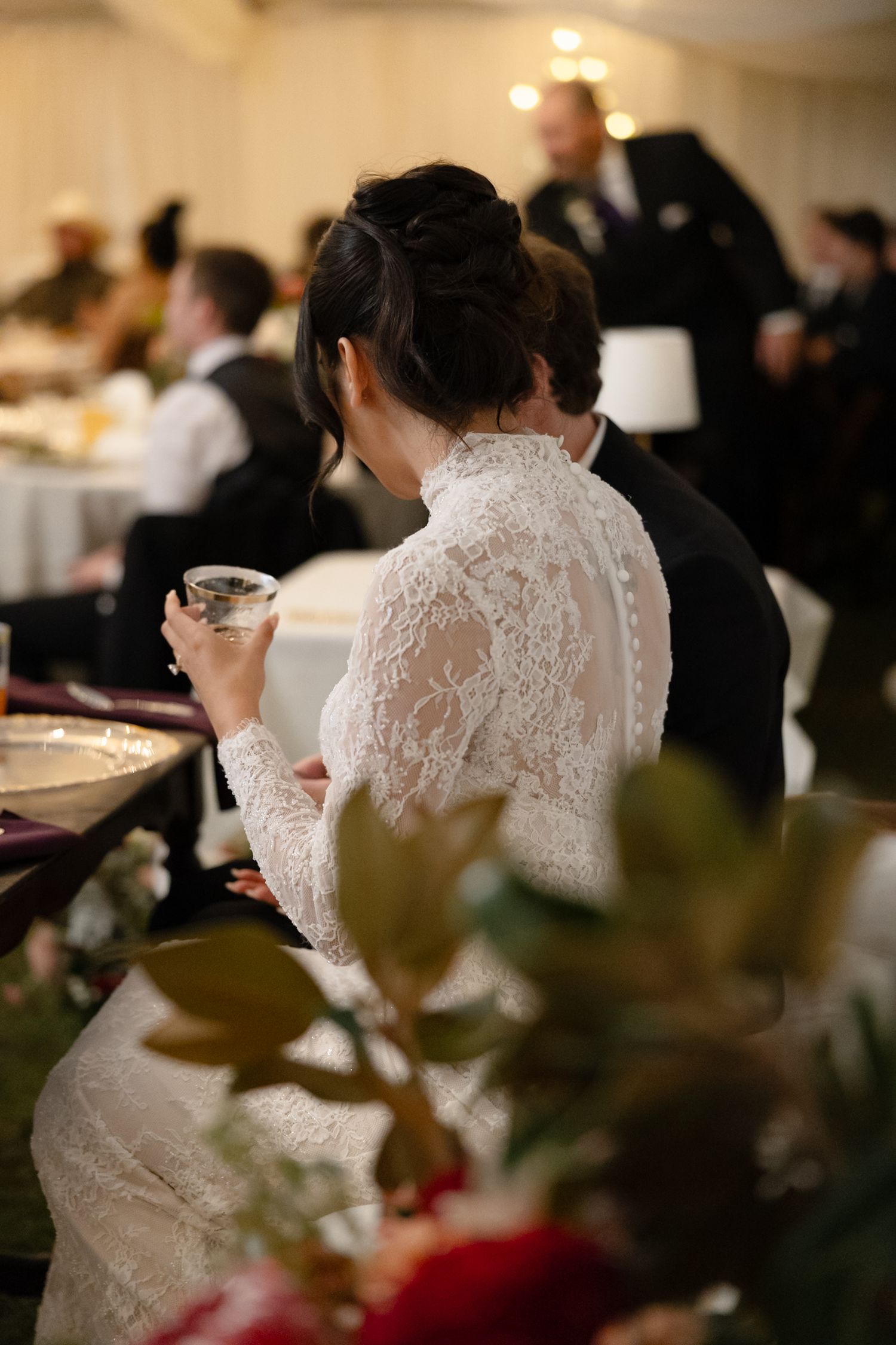Bride in long-sleeve lace wedding gown seated at reception table holding a glass during wedding celebration.