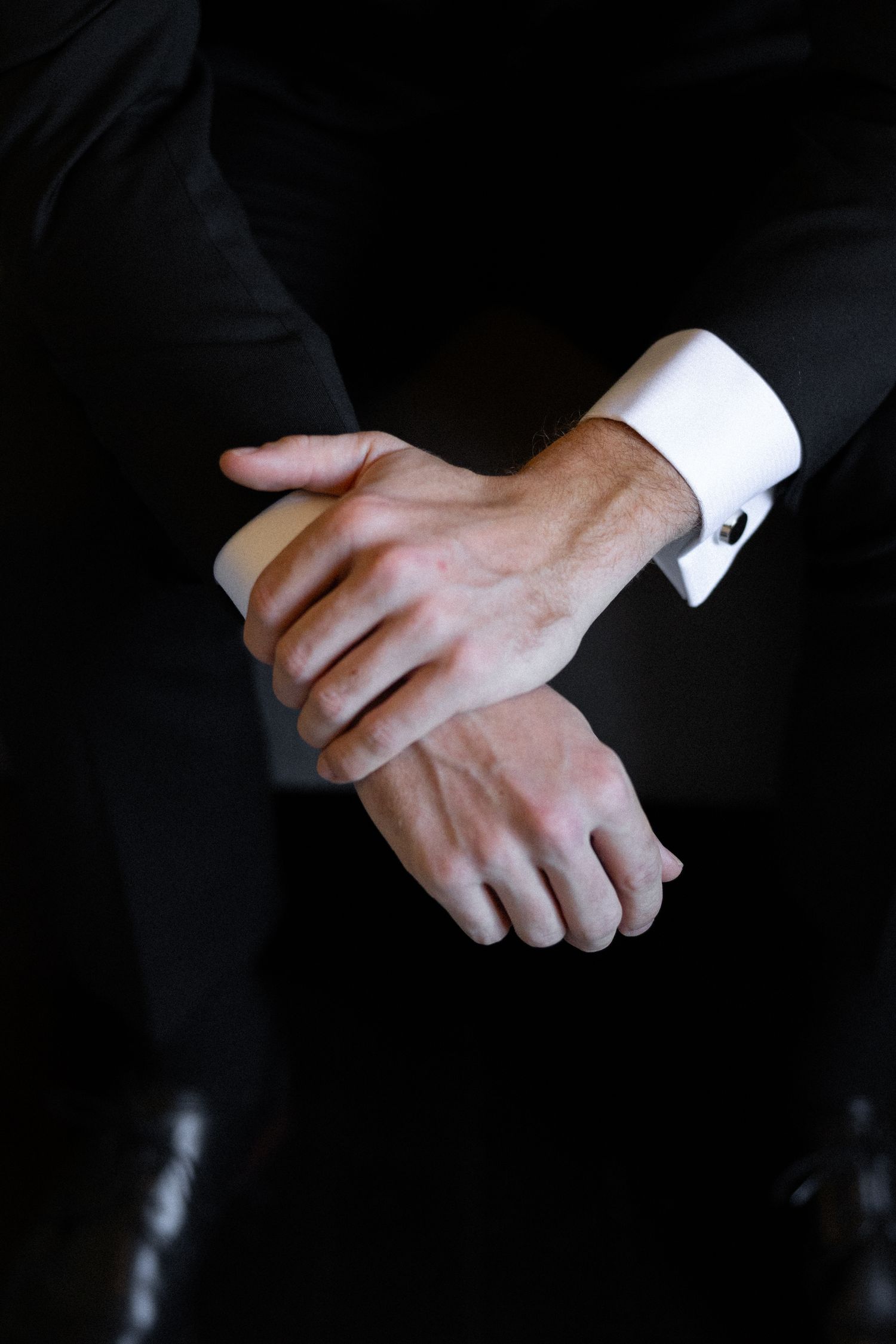 Close-up of a man in a black tuxedo with white French cuff shirt, hands clasped together.