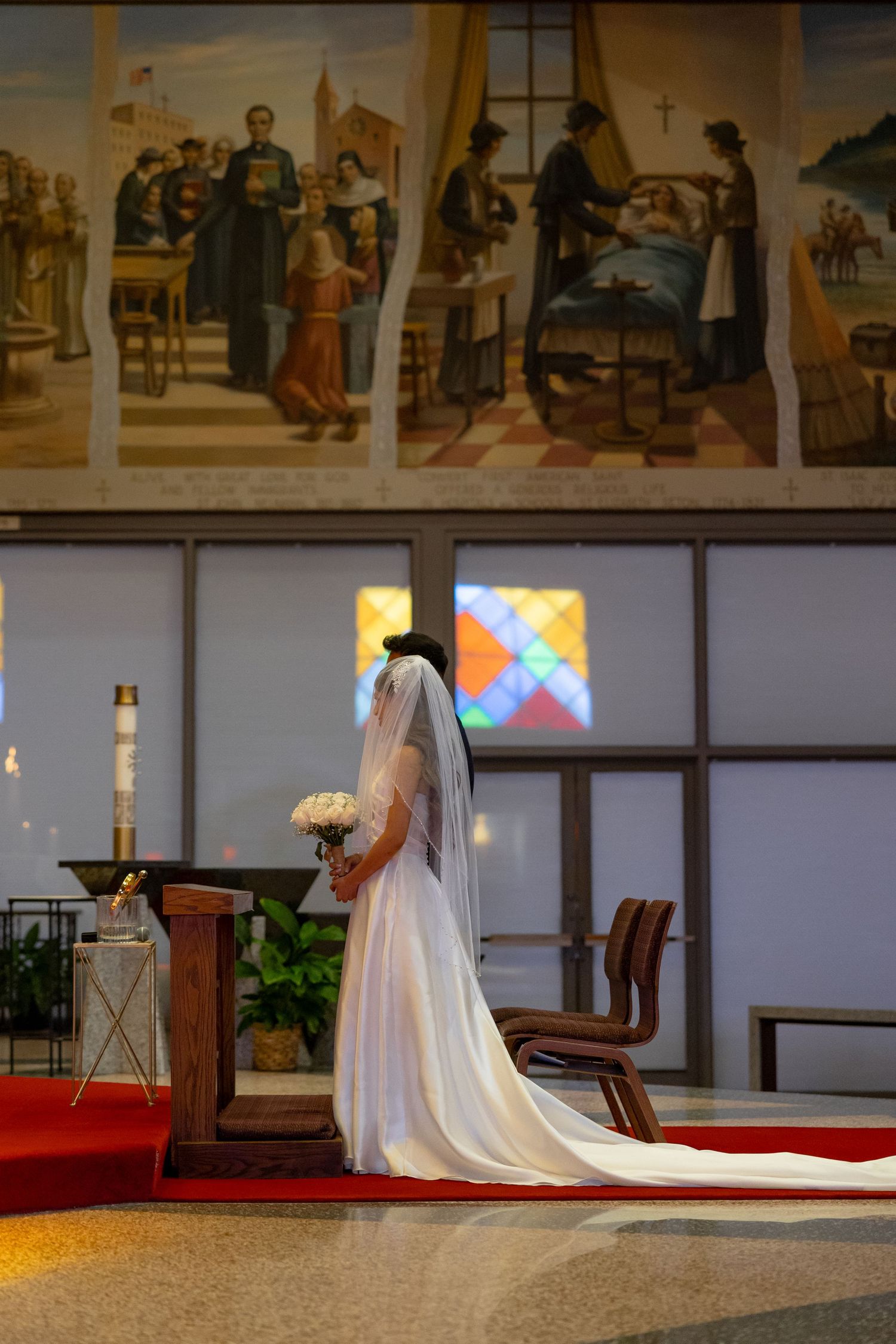 Bride in white gown and veil kneeling at church altar with stained glass windows and religious murals.