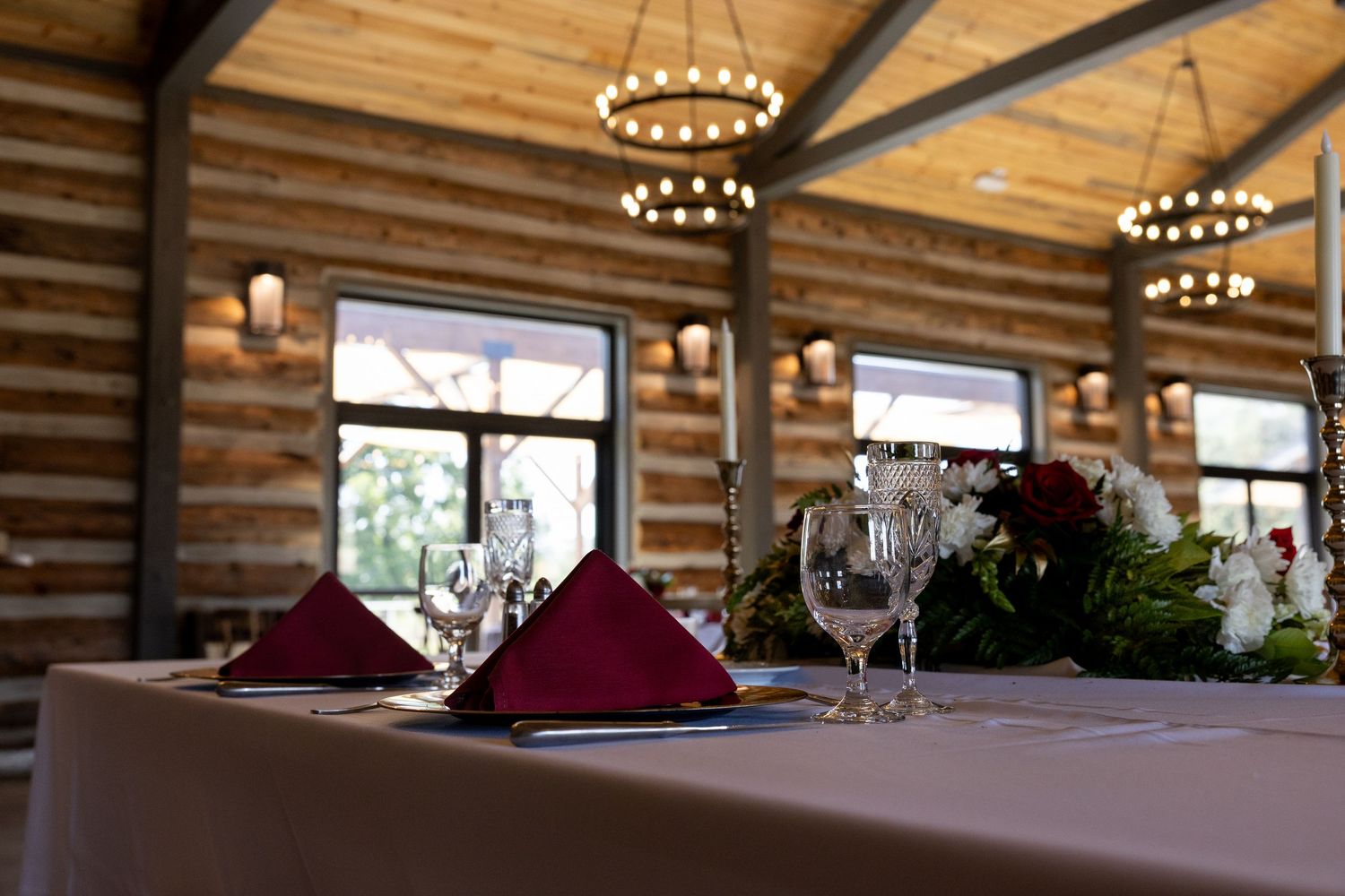 Elegant log cabin dining room with crimson napkins, crystal glasses, and floral centerpieces under chandelier lighting.