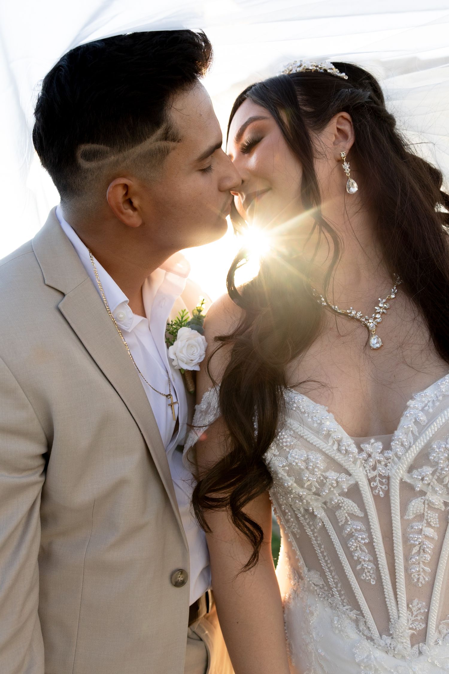 Bride and groom share a romantic sunlit kiss, bride wearing lace gown and crystal jewelry on wedding day.