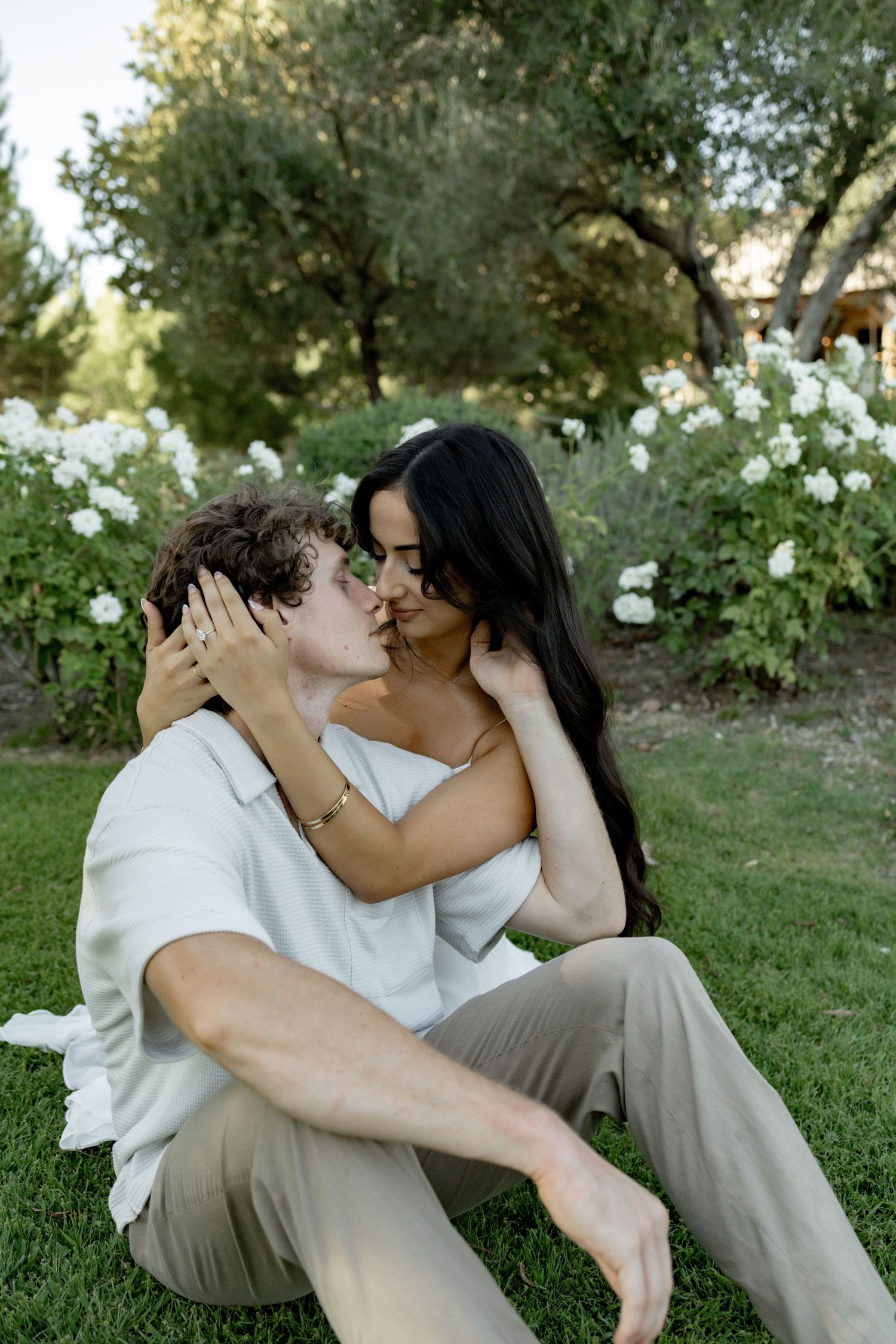 A couple shares intimate moments while sitting together on grass with white flowers and trees in the background.