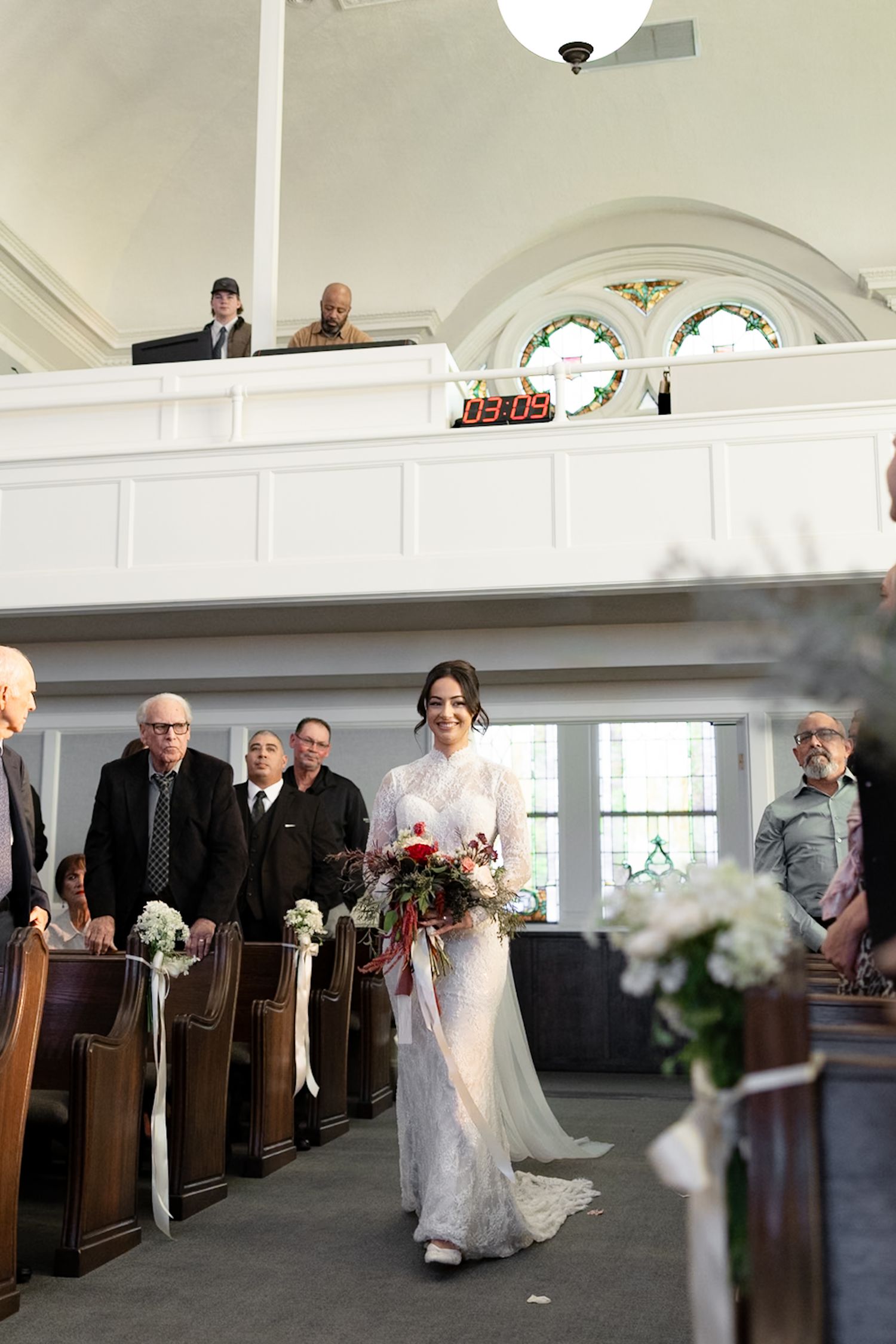 Bride walks down church aisle in lace gown holding red and white bouquet as guests stand in wooden pews.