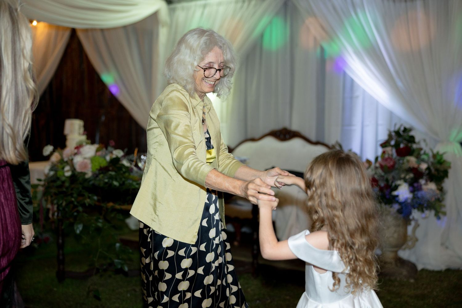 Elderly woman in green jacket dances with young girl in white dress at wedding reception.