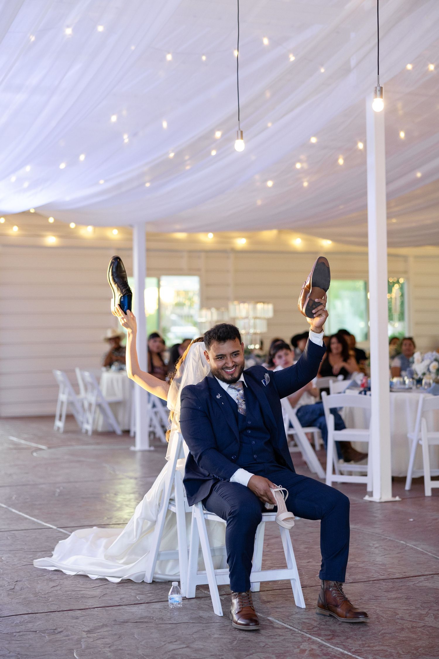 Groom in navy suit holds up shoes while seated with bride during fun wedding reception game.