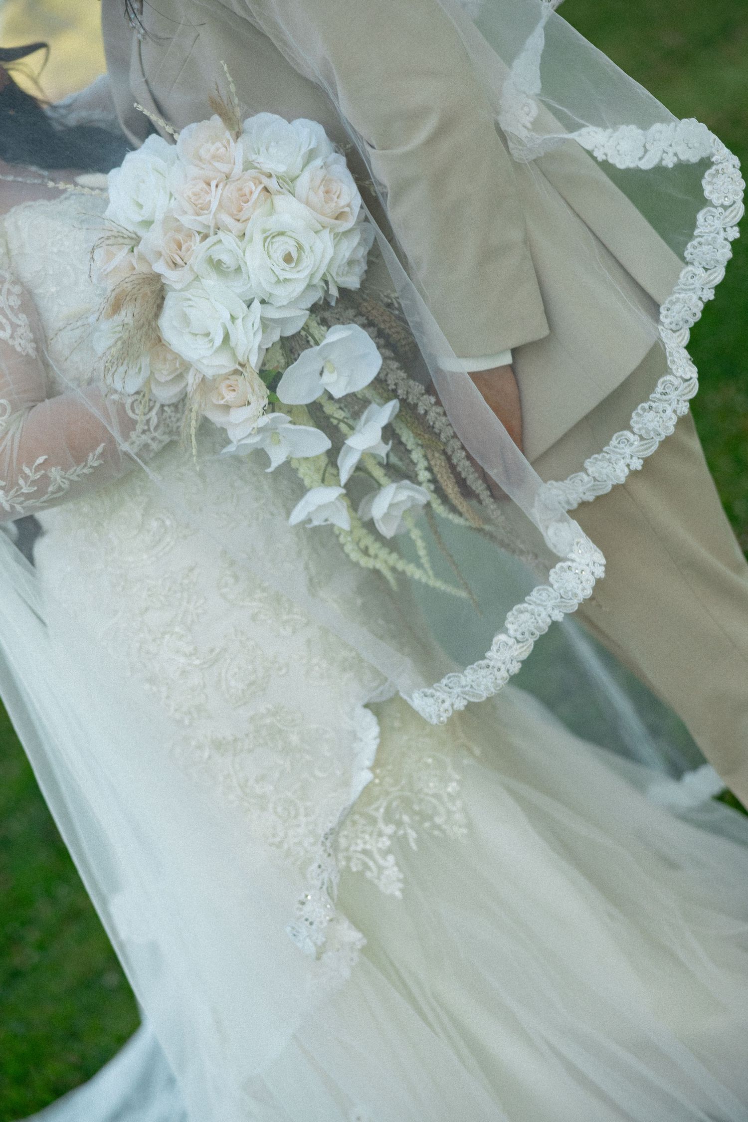 Bride in lace wedding gown holding white rose bouquet with groom in tan suit outdoors.