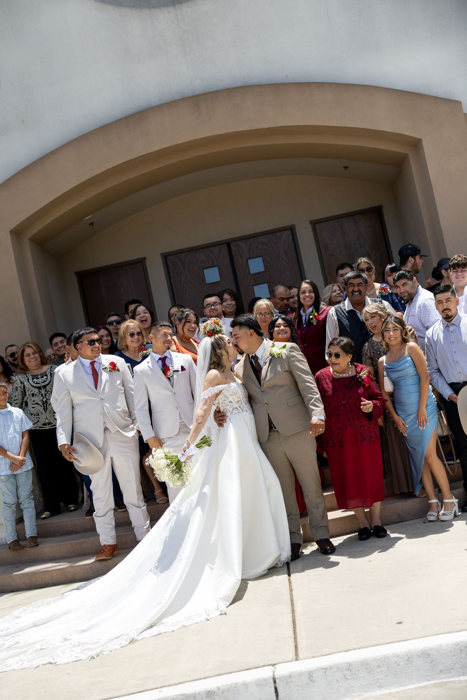 Bride and groom share a kiss on church steps surrounded by wedding guests in formal attire on a sunny day.
