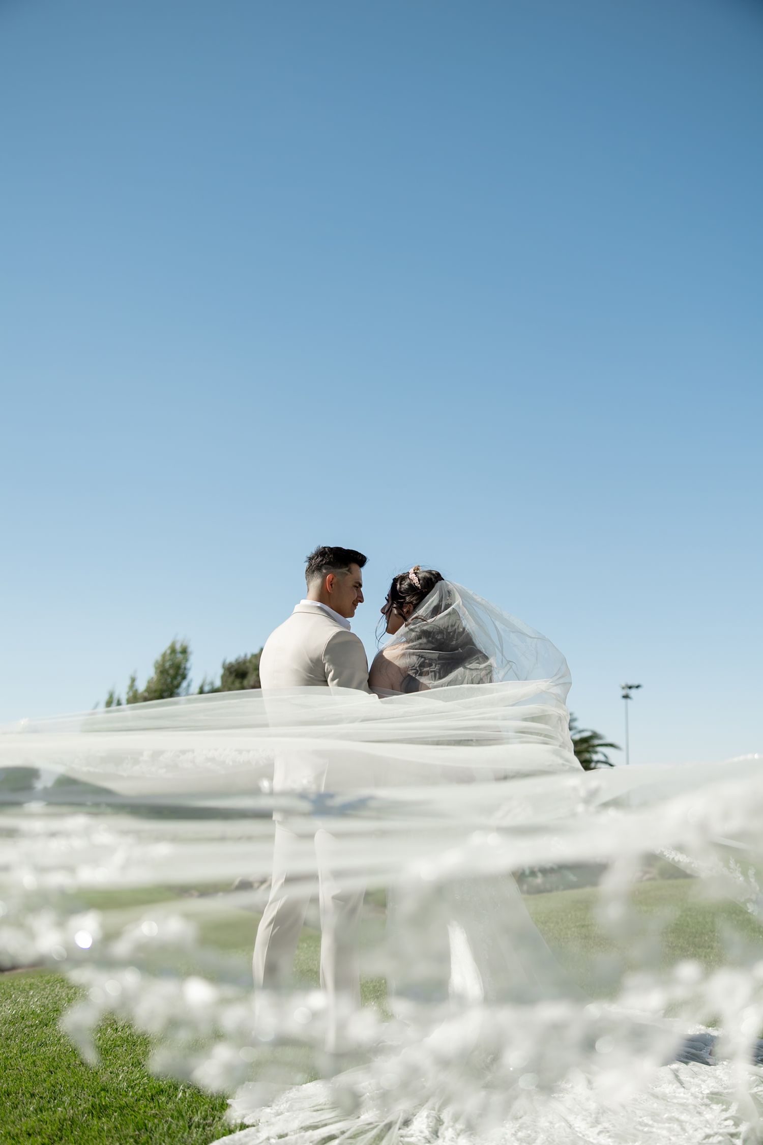Bride and groom face each other outdoors under blue sky, with flowing white veil billowing in the foreground.
