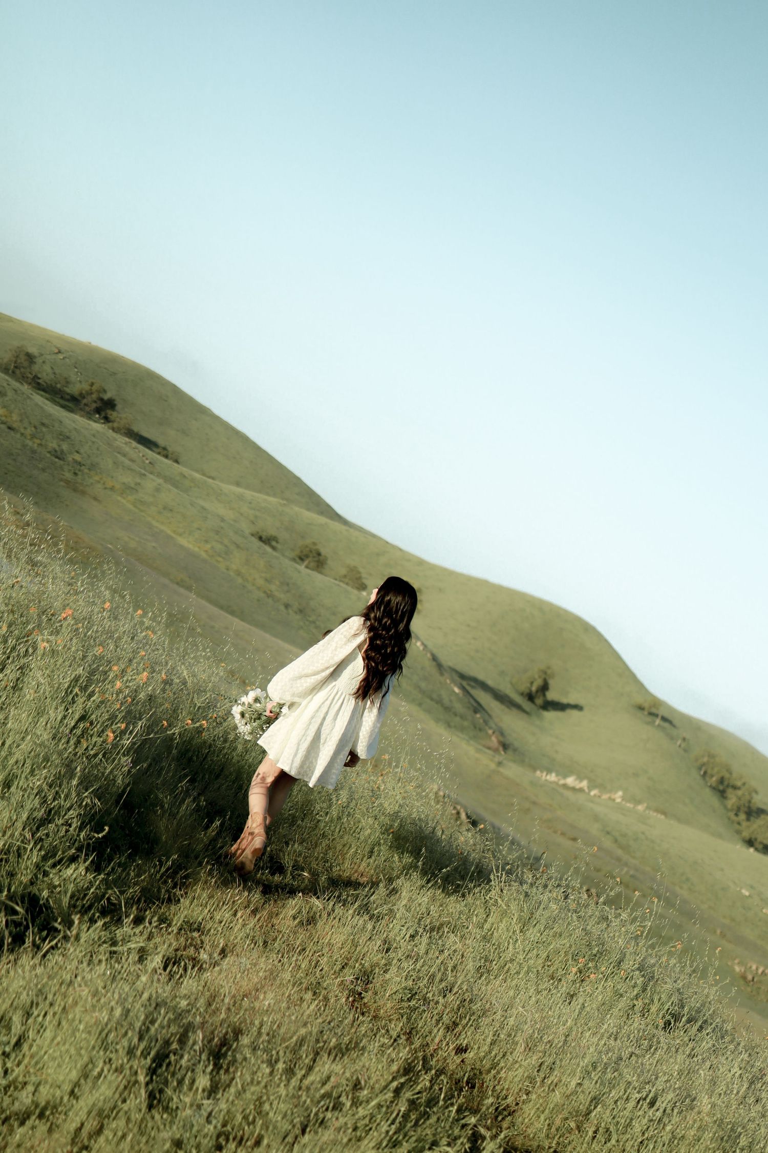 Person in white dress walking through tall grass on a hillside.