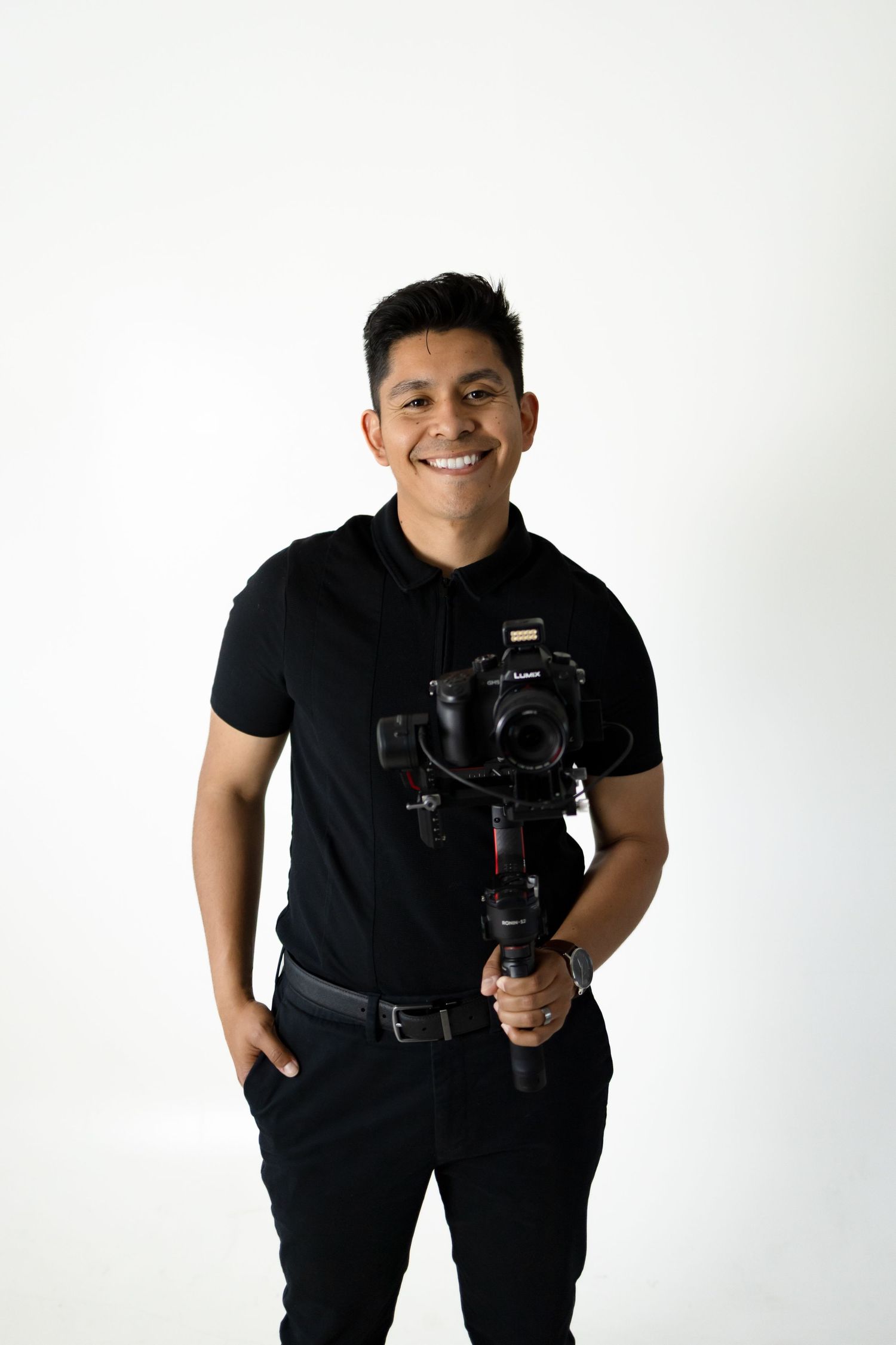 Professional photographer in all black attire holding a camera gimbal setup against white background.