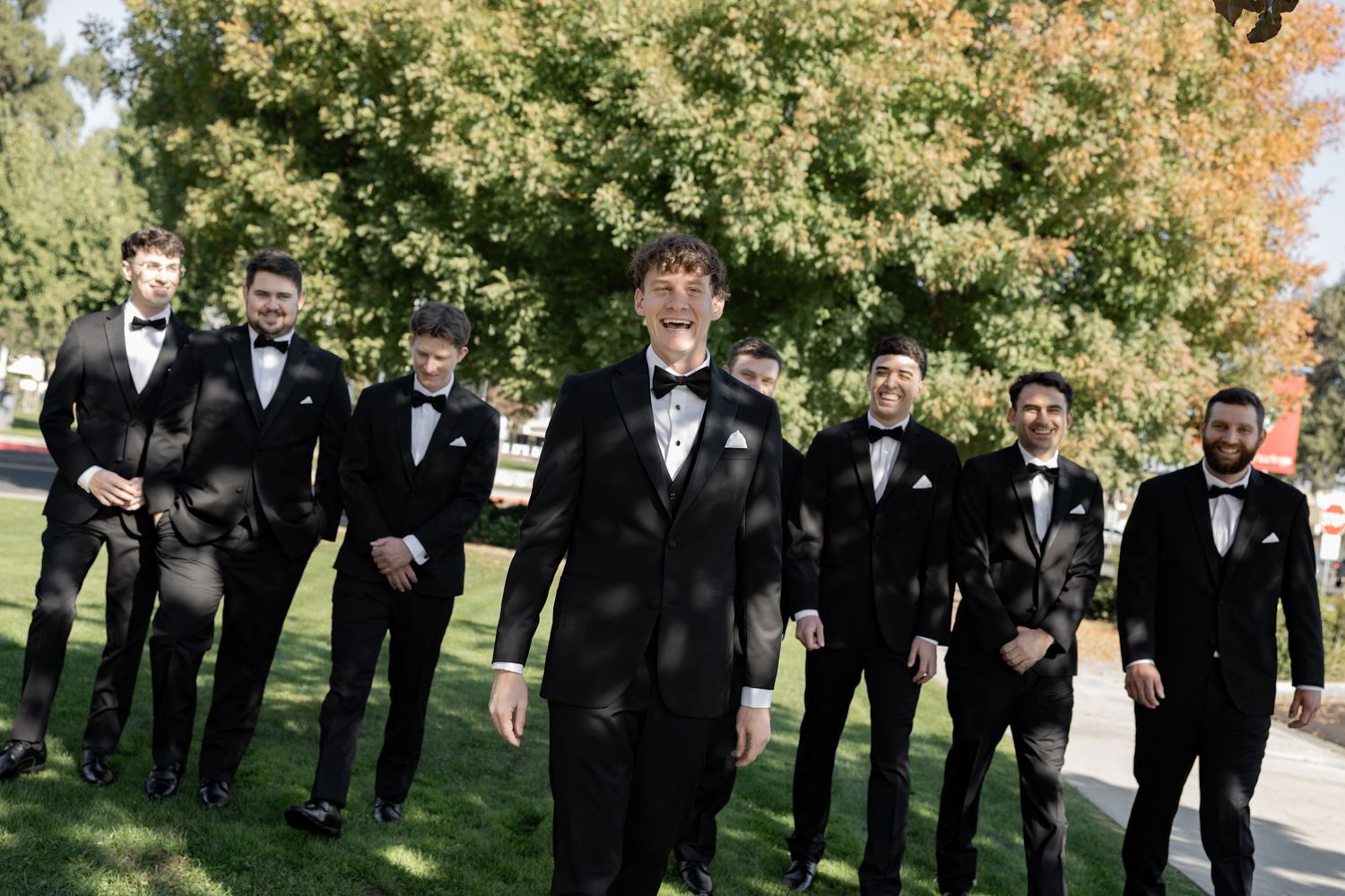 Groom and groomsmen in black tuxedos with bow ties posing outdoors on a sunny day at a wedding.