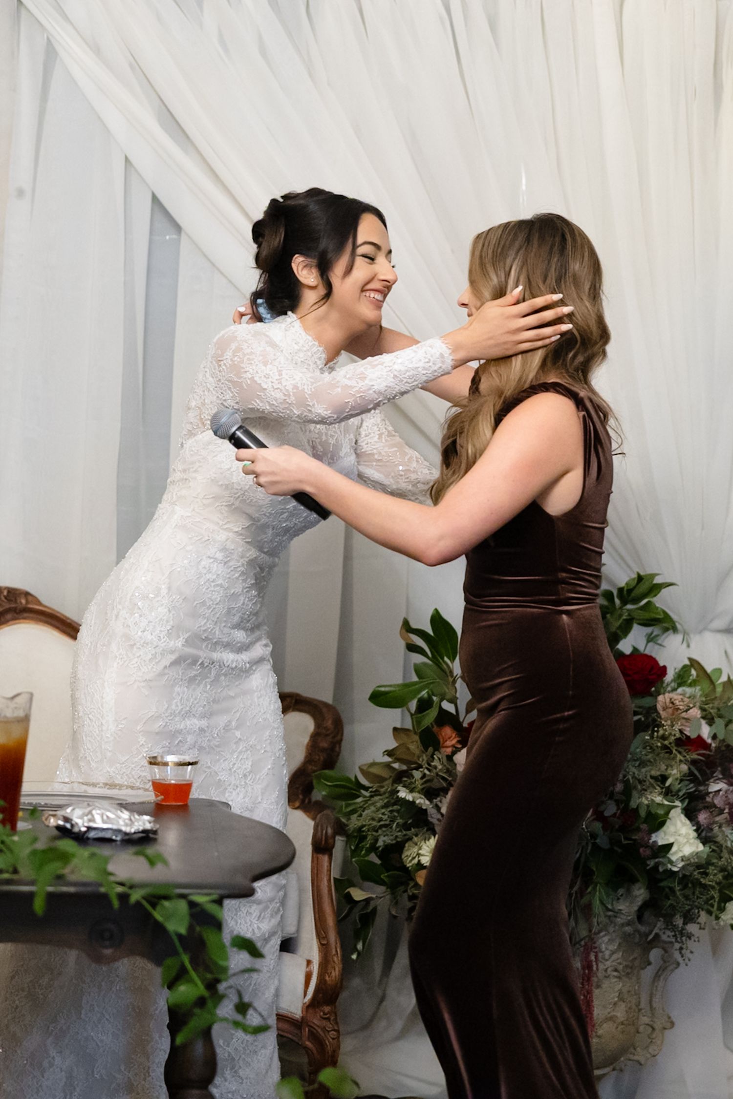Bride in white lace gown embraces woman in brown velvet dress at wedding reception.