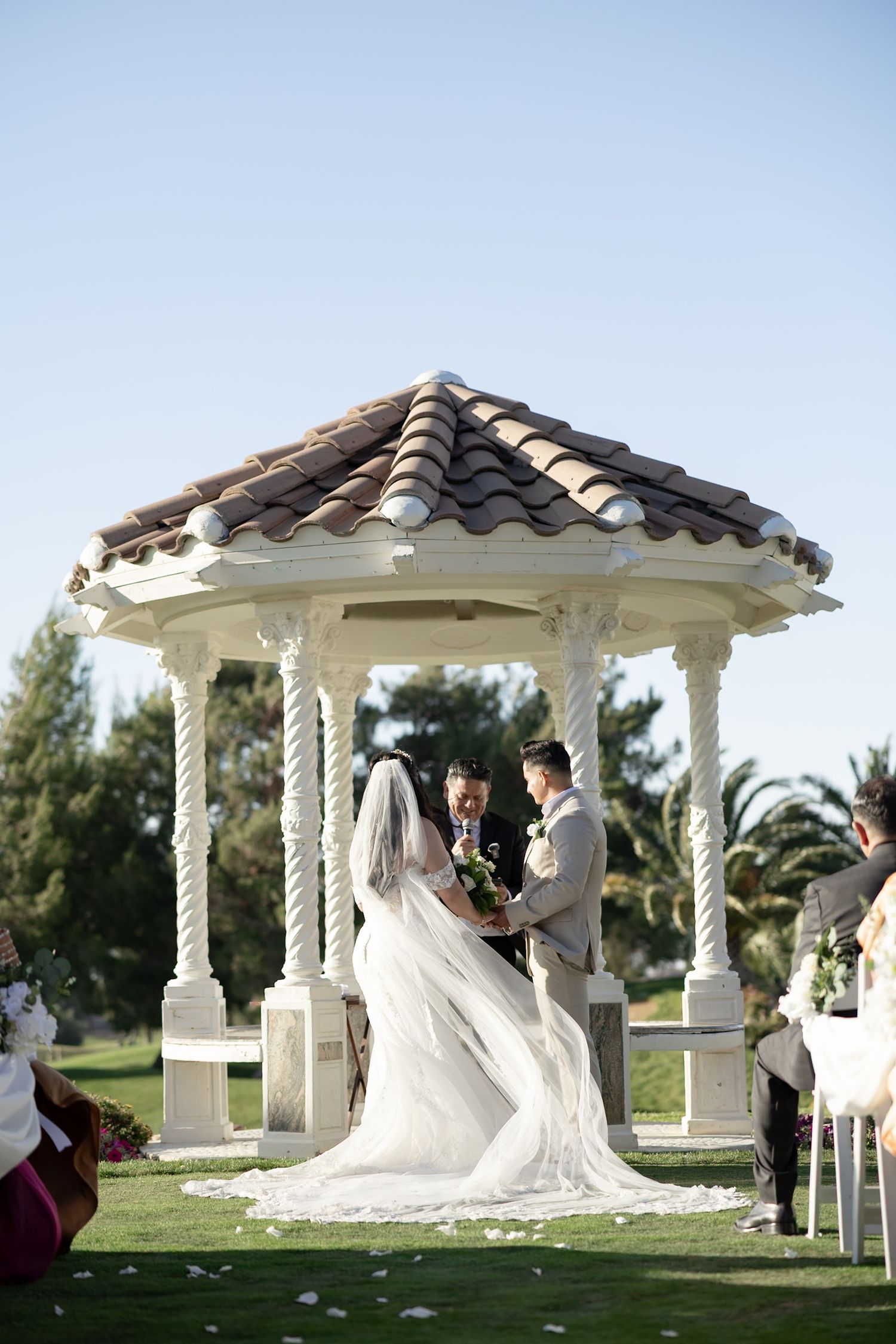 Bride and groom exchange vows under elegant white gazebo with terracotta roof on lush green lawn.