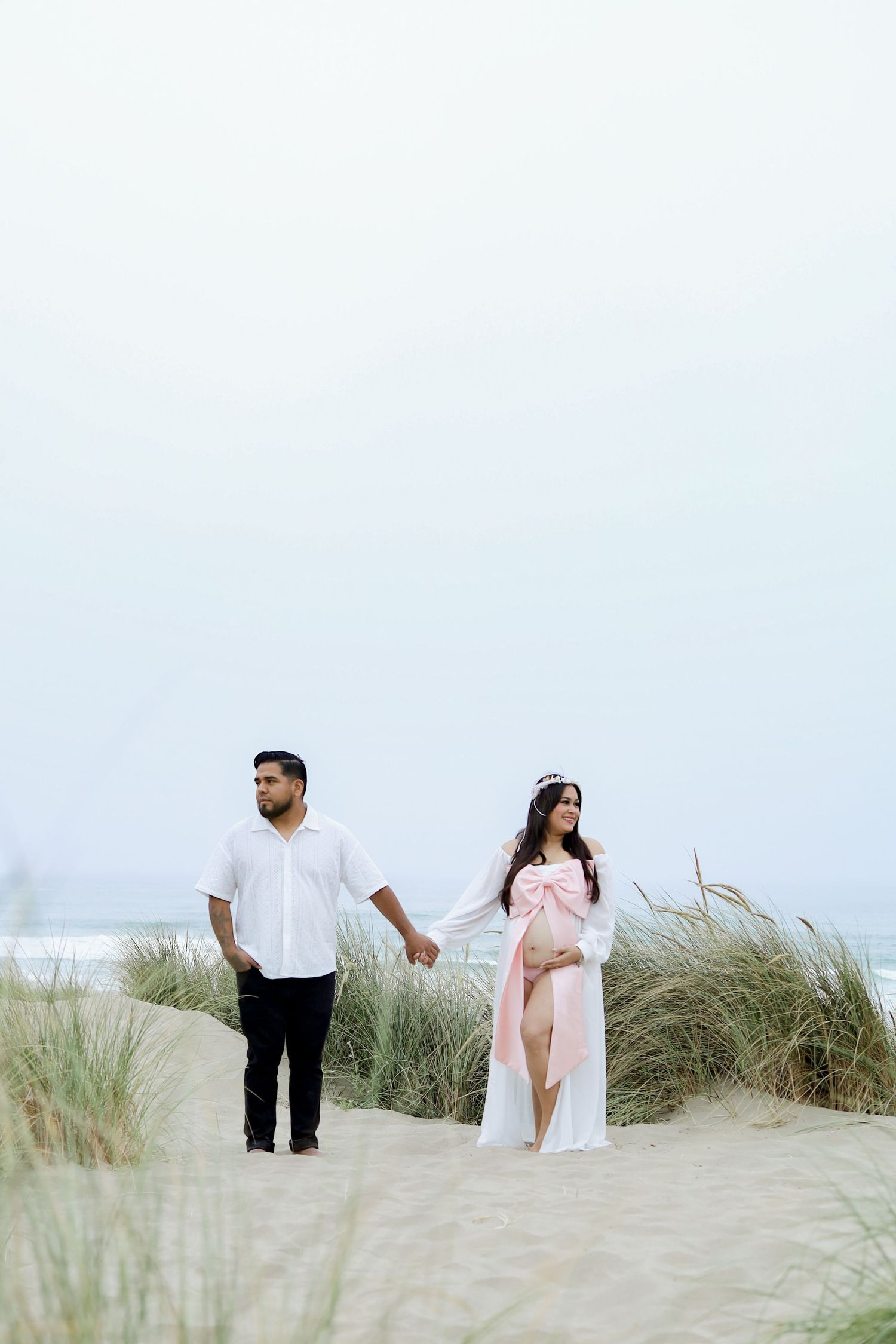 A couple walking together on sandy dunes with tall grass against a bright white sky.