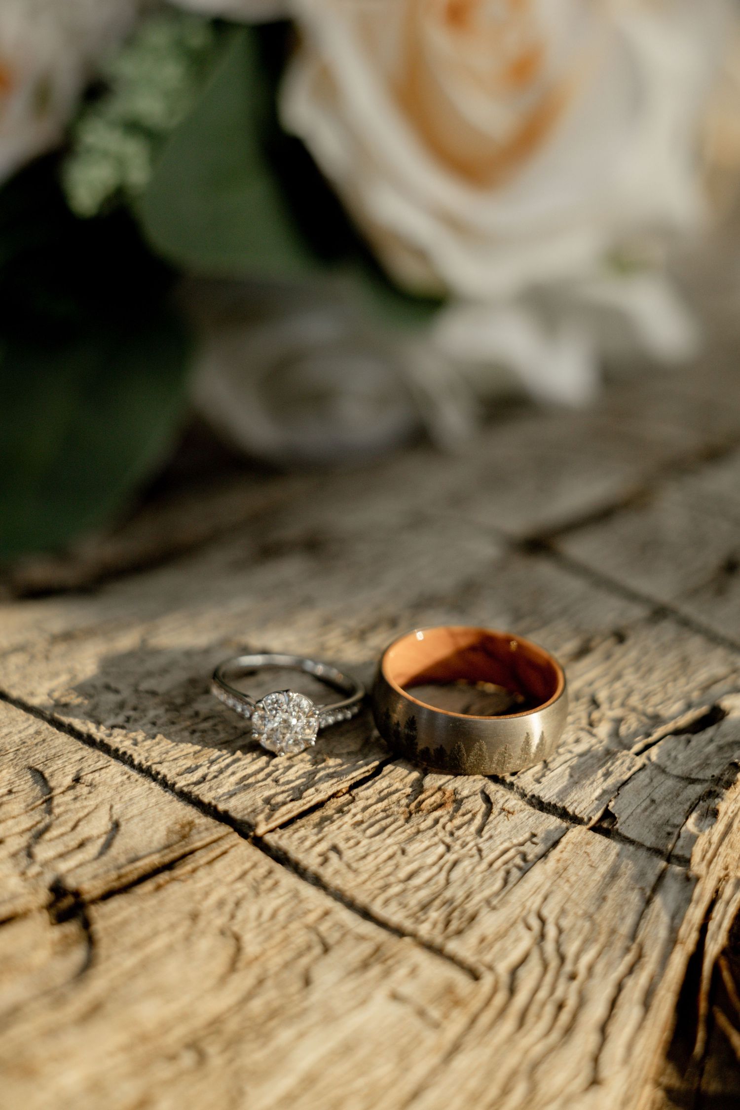 Wedding rings and diamond engagement ring artistically photographed on rustic wooden surface with roses in background.