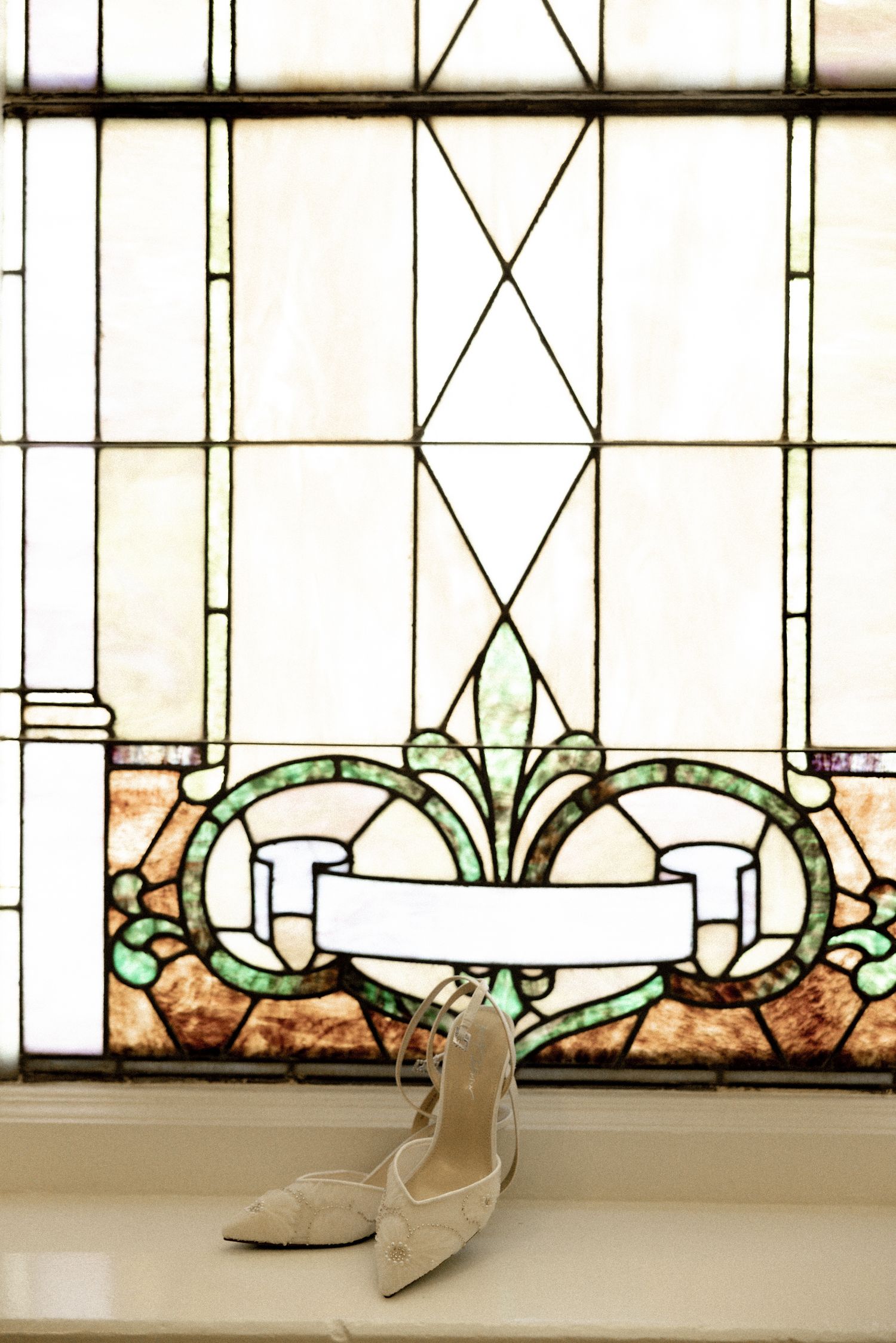 Elegant bridal heels displayed on a windowsill in front of a decorative stained glass window.