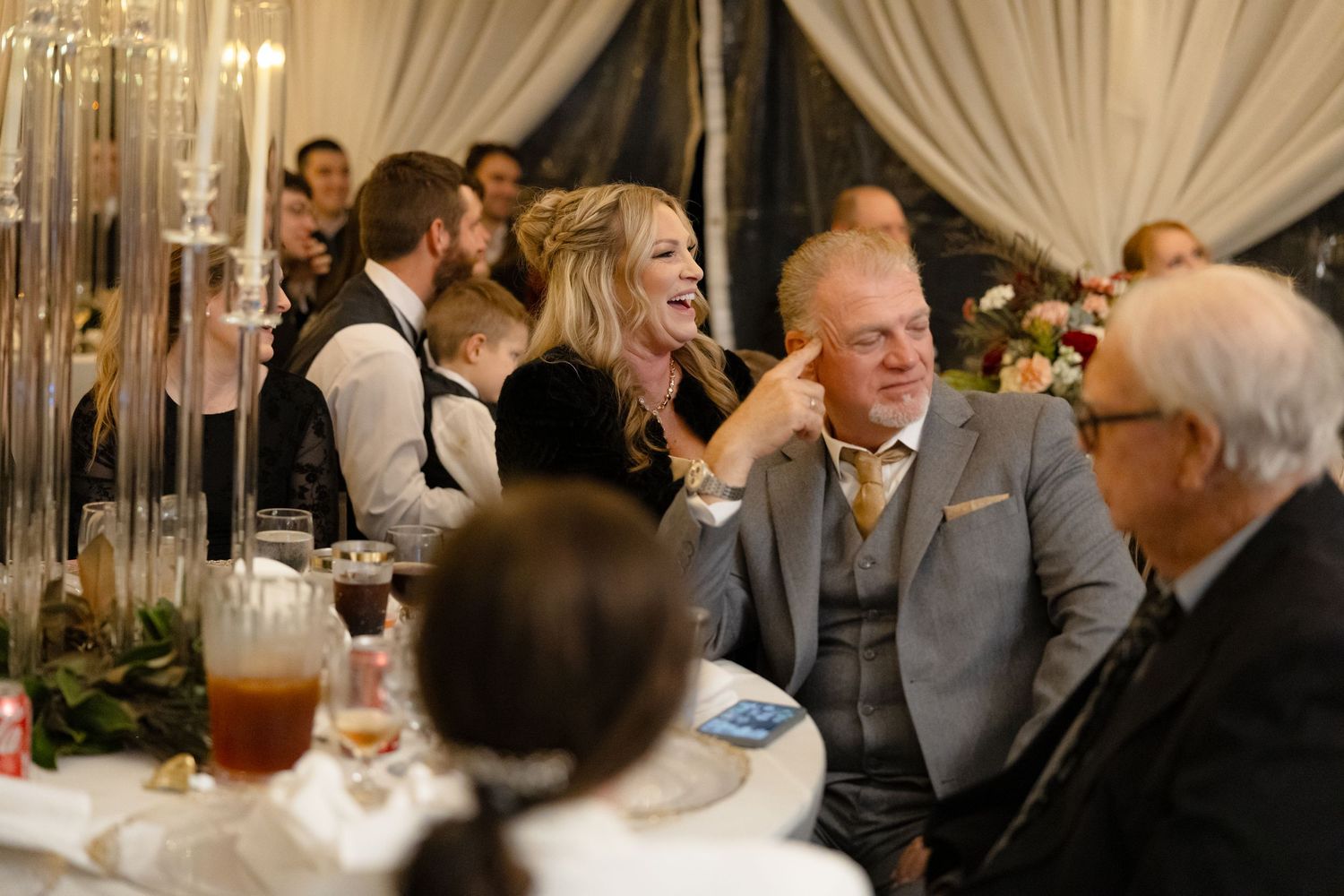 Wedding guests laughing and celebrating at elegant reception dinner under decorated tent.