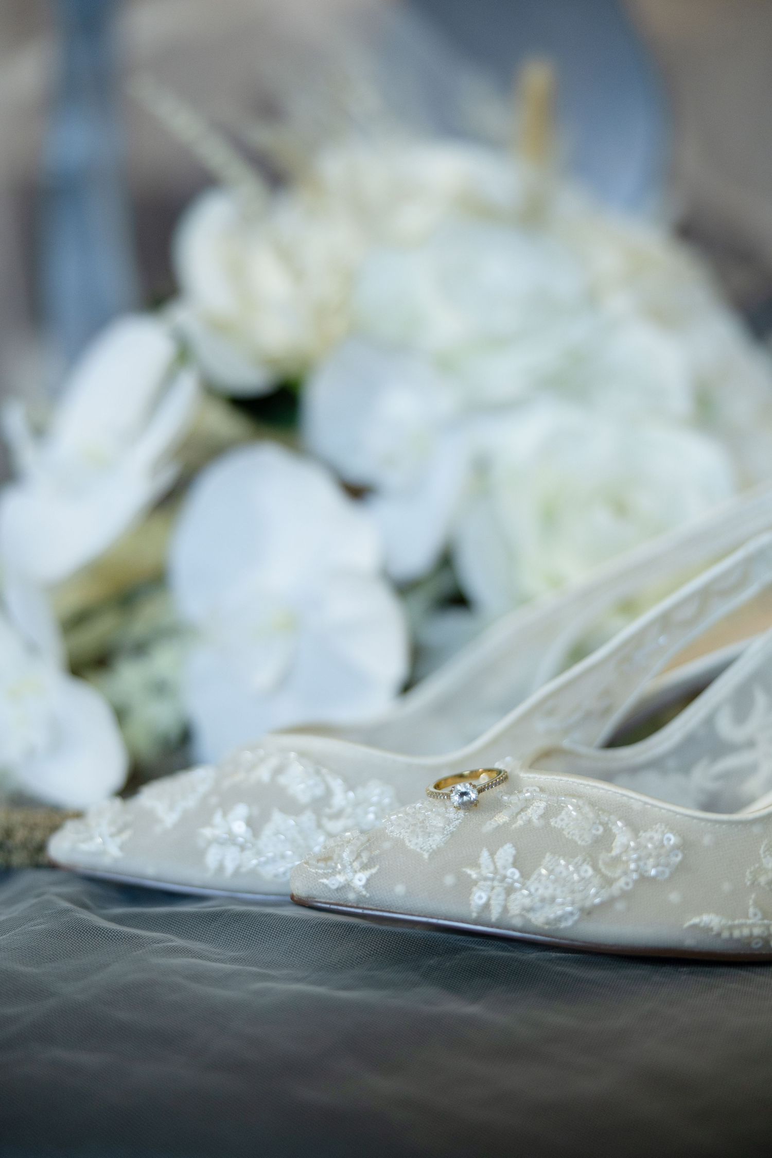 Elegant white lace bridal heels with a gold diamond ring resting on top, beside a bouquet of white orchids and roses.