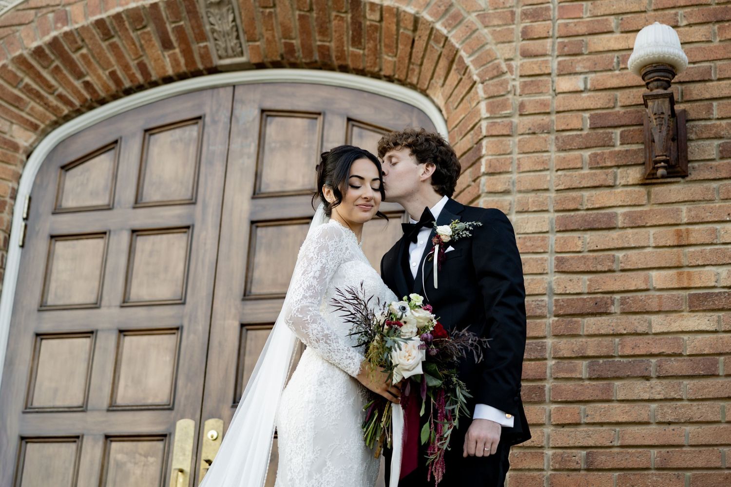 Bride and groom sharing a kiss outside brick church with arched wooden doors.