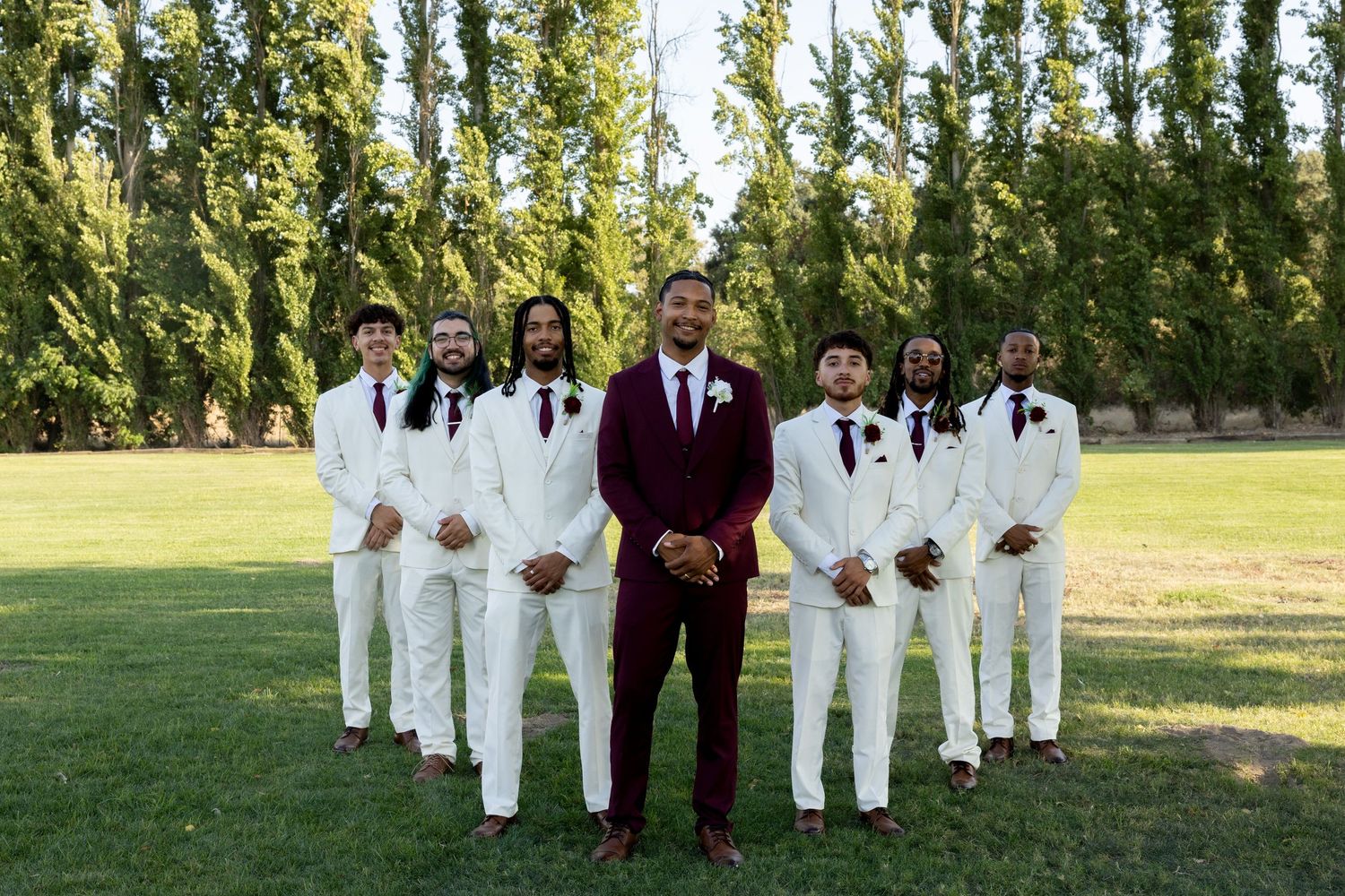 Groom in burgundy suit stands with six groomsmen in white suits on green lawn with tall trees backdrop.