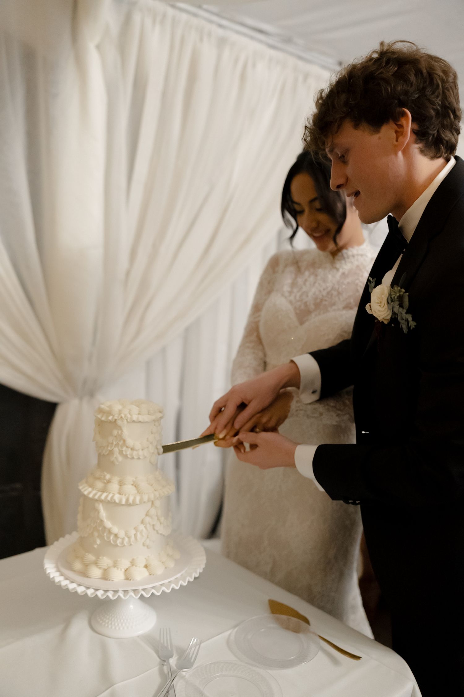Bride and groom smiling while cutting a three-tier white wedding cake together.