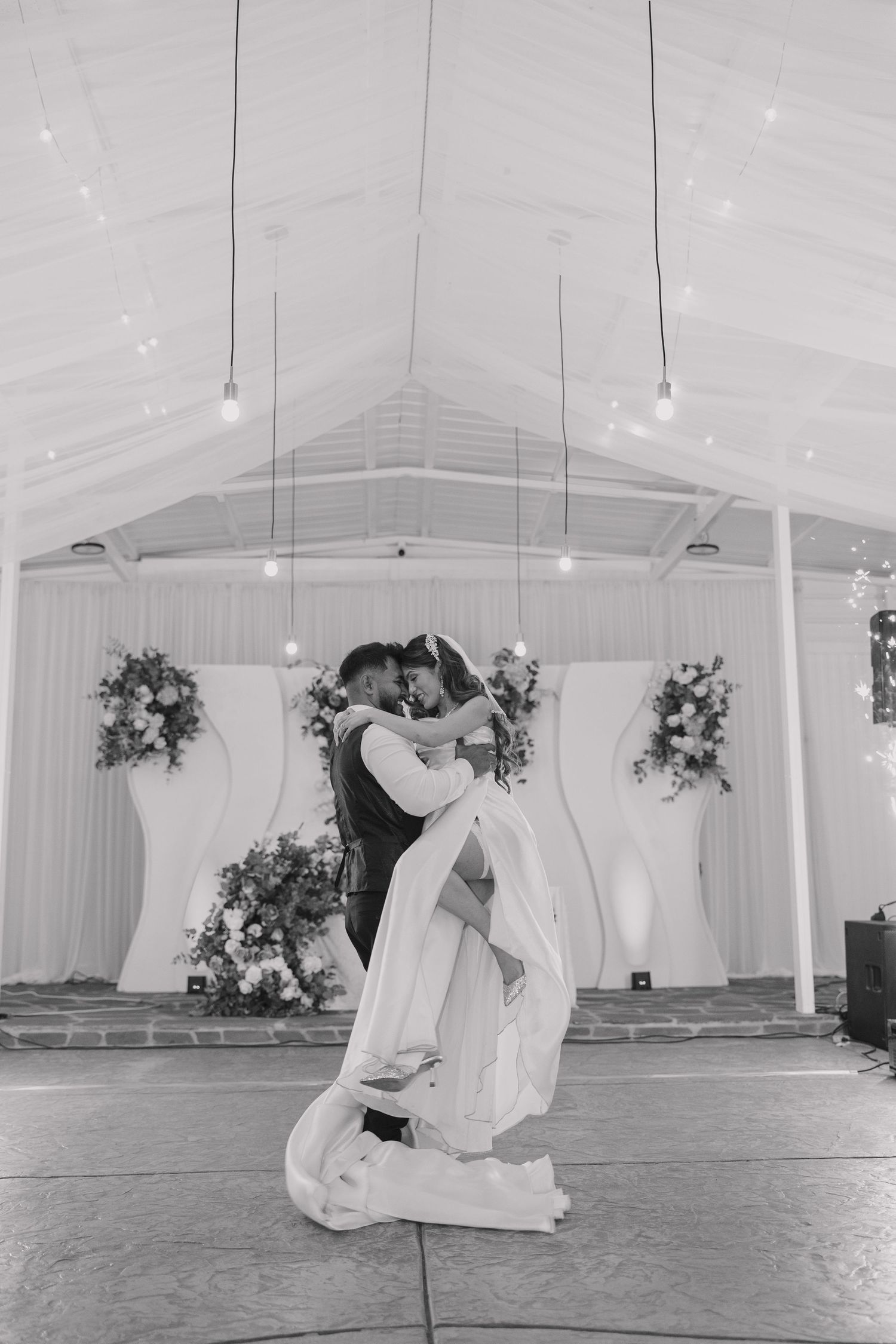 Black and white photo of a groom lifting his bride during their first dance in an elegant decorated wedding venue.