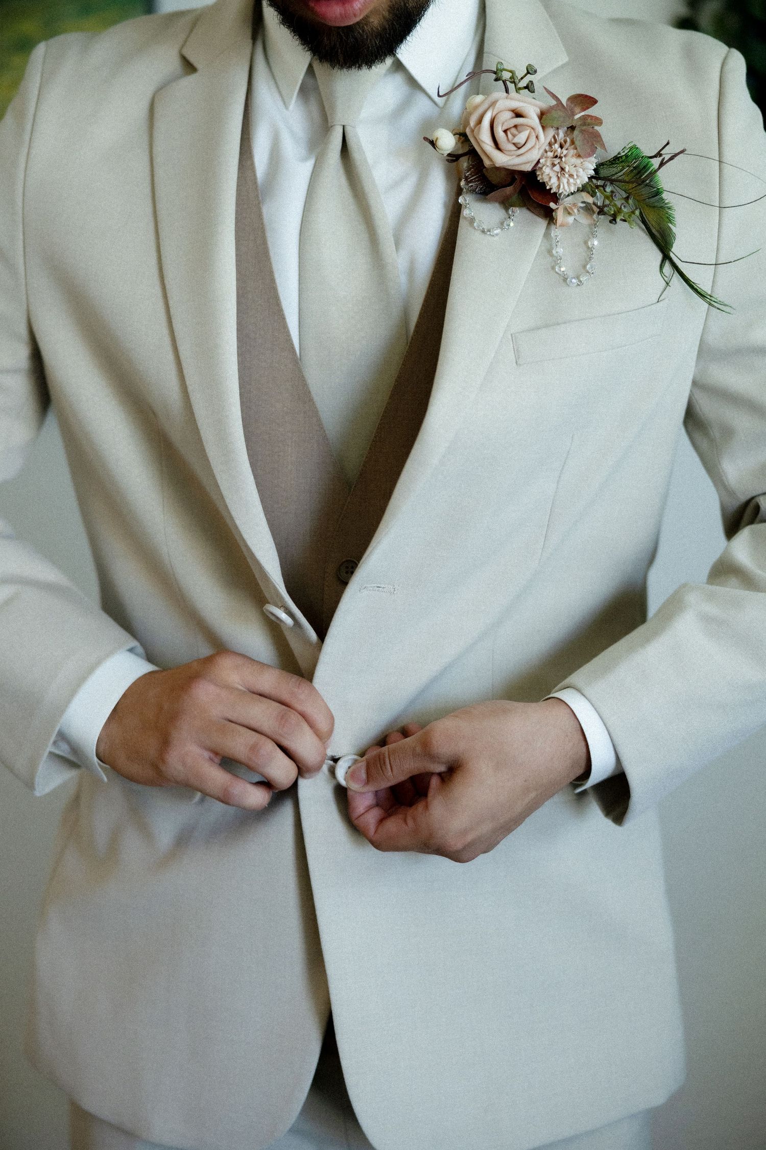 Groom in beige suit buttoning jacket with floral boutonniere featuring blush roses on wedding day.