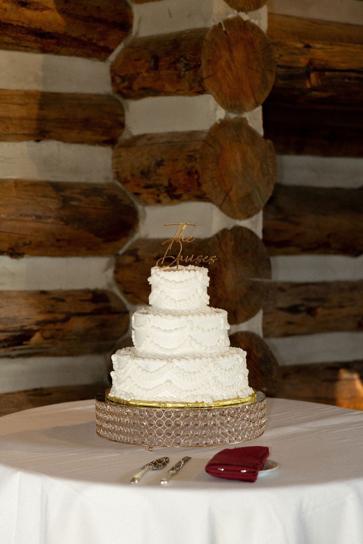 Elegant three-tier white wedding cake with gold topper on crystal stand against rustic log cabin wall.