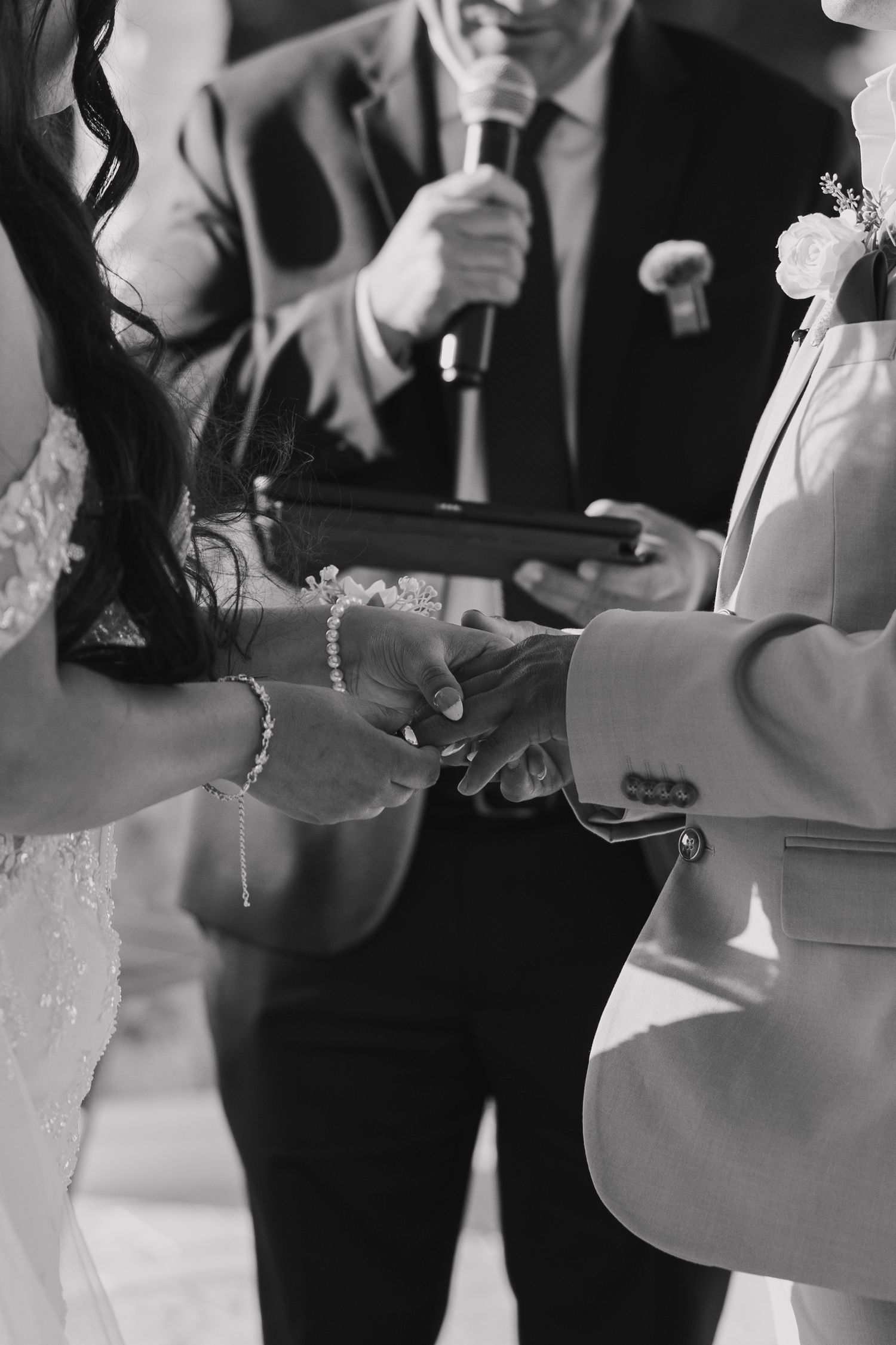 Black and white photo of bride and groom exchanging rings during wedding ceremony with officiant holding microphone.